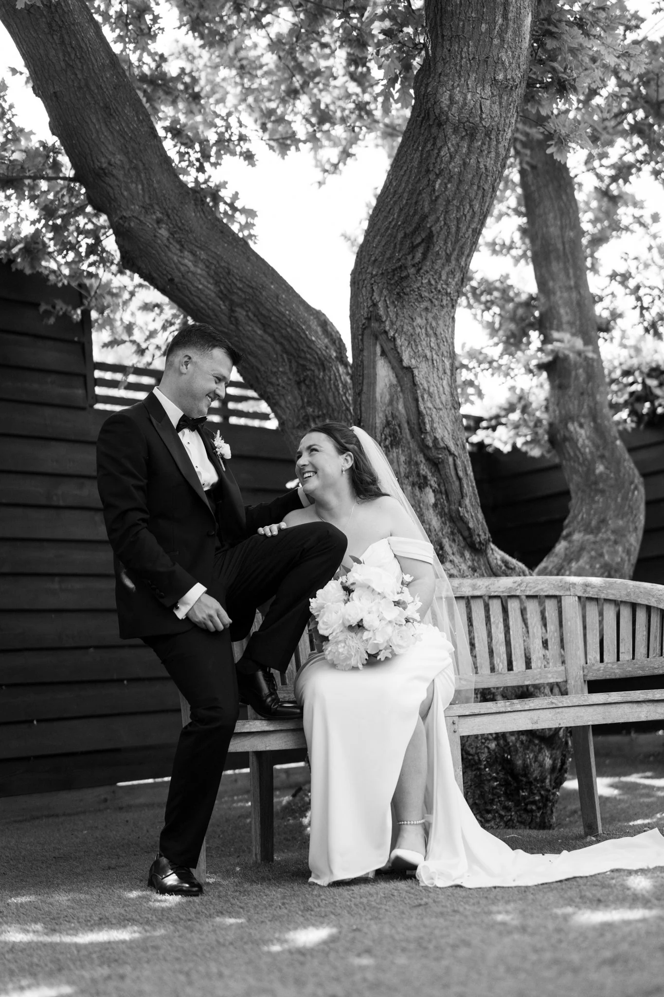 A black and white photo of a wedding couple outdoors. The groom, dressed in a tuxedo, is smiling and sitting on a wooden bench while the bride, wearing a long wedding dress and veil, is smiling and looking up at him. They are surrounded by large tree