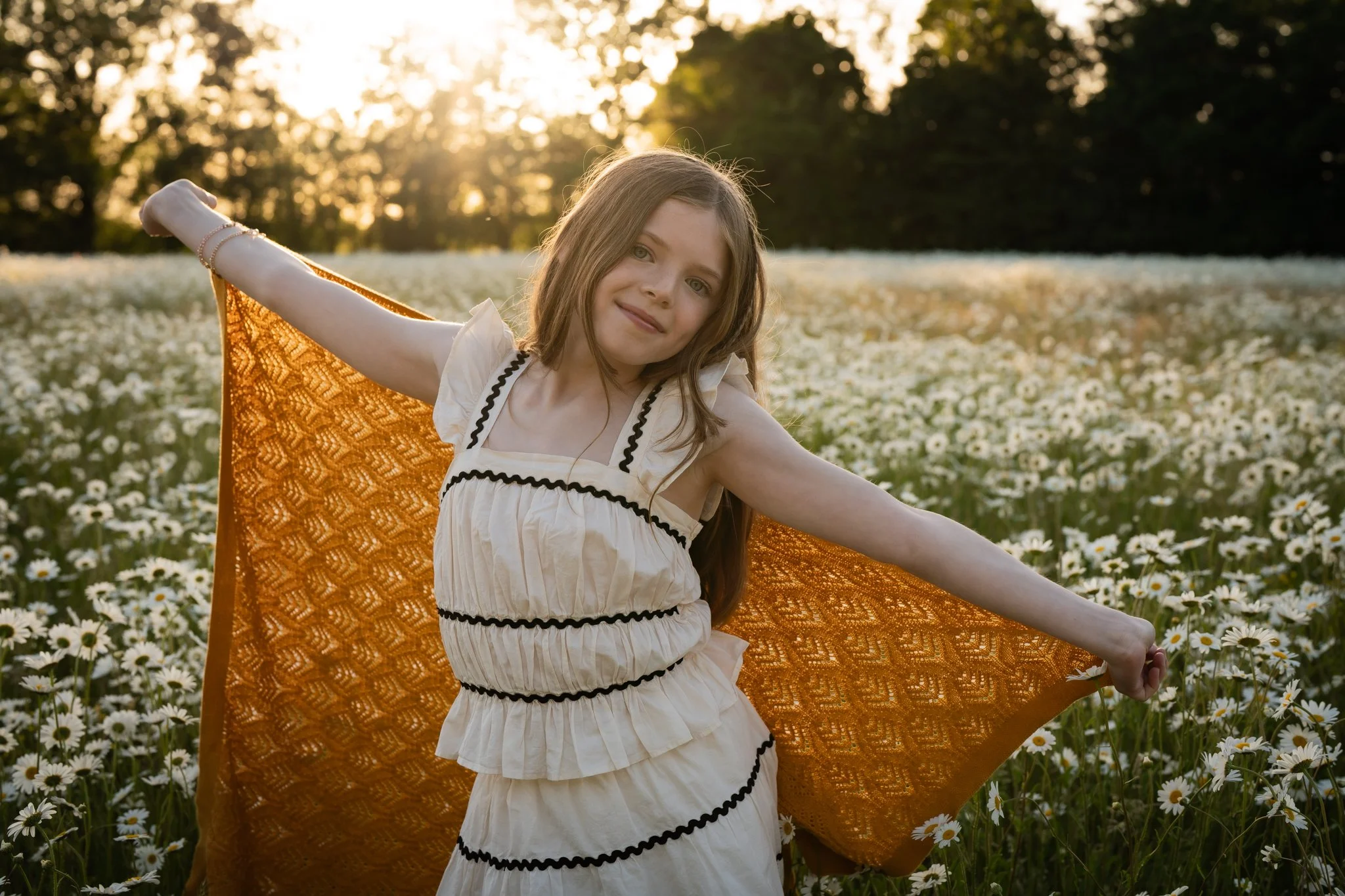 A girl standing in a field of white daisies holding an orange fabric. The setting sun is behind her, creating a backlit scene with warm light. Anne Wills Photography. Braintree, Essex