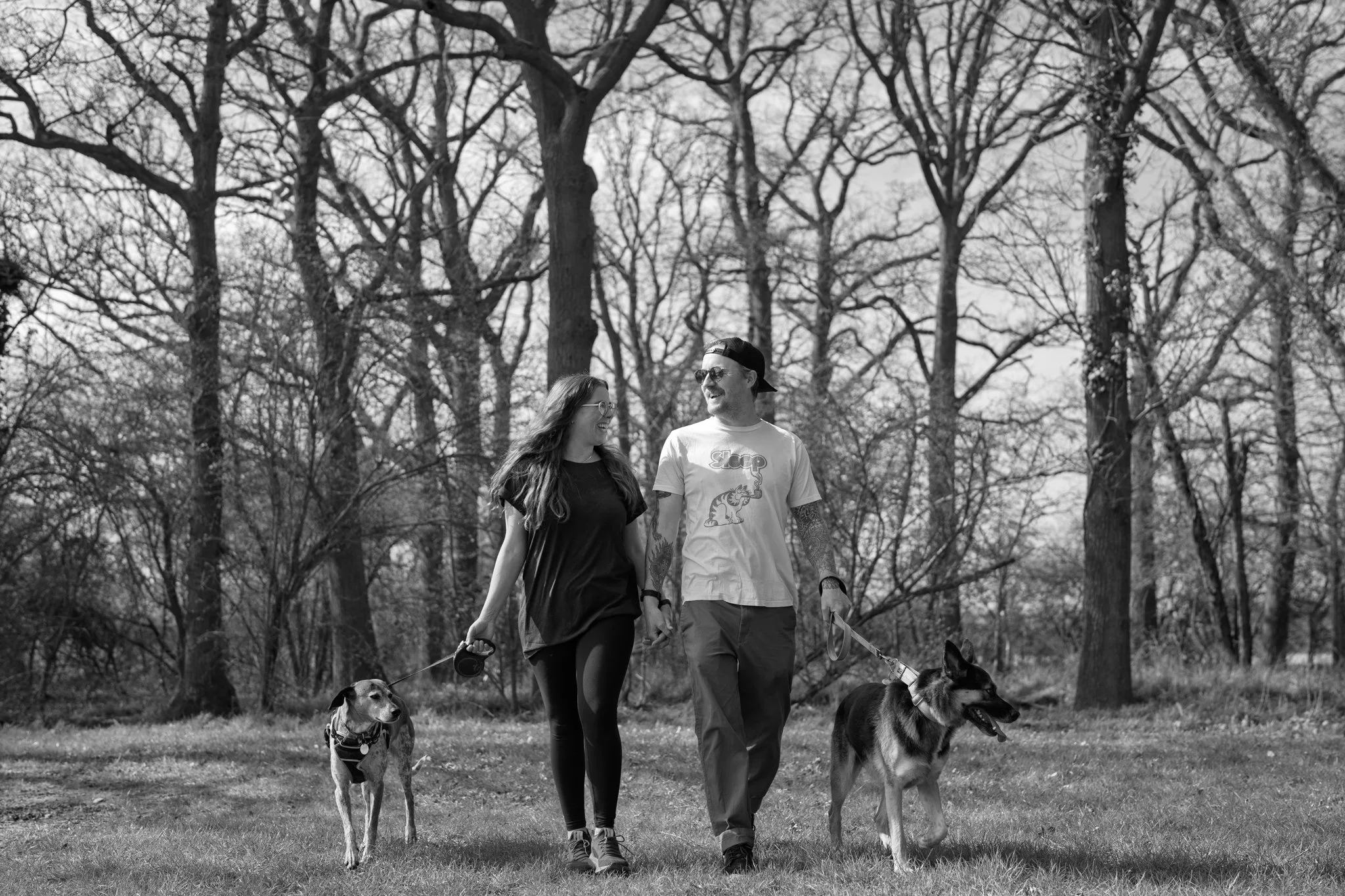 A black and white photograph of two people walking their dogs in a park with leafless trees in the background. The man is wearing a baseball cap backwards, sunglasses, and a T-shirt, while the woman has long hair, glasses, and a dark T-shirt. They ar