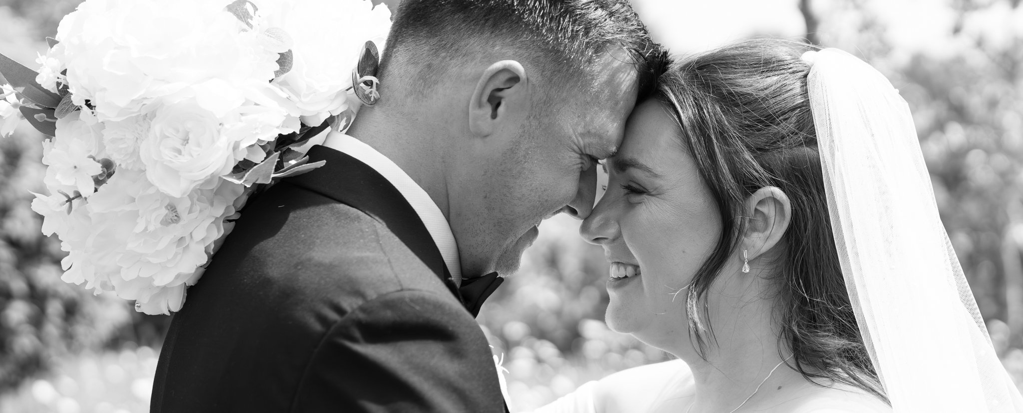 Black and white photograph of a bride and groom touching foreheads, smiling, outdoors, with flowers and trees in the background. Anne Wills Photography Maidens barn Essex