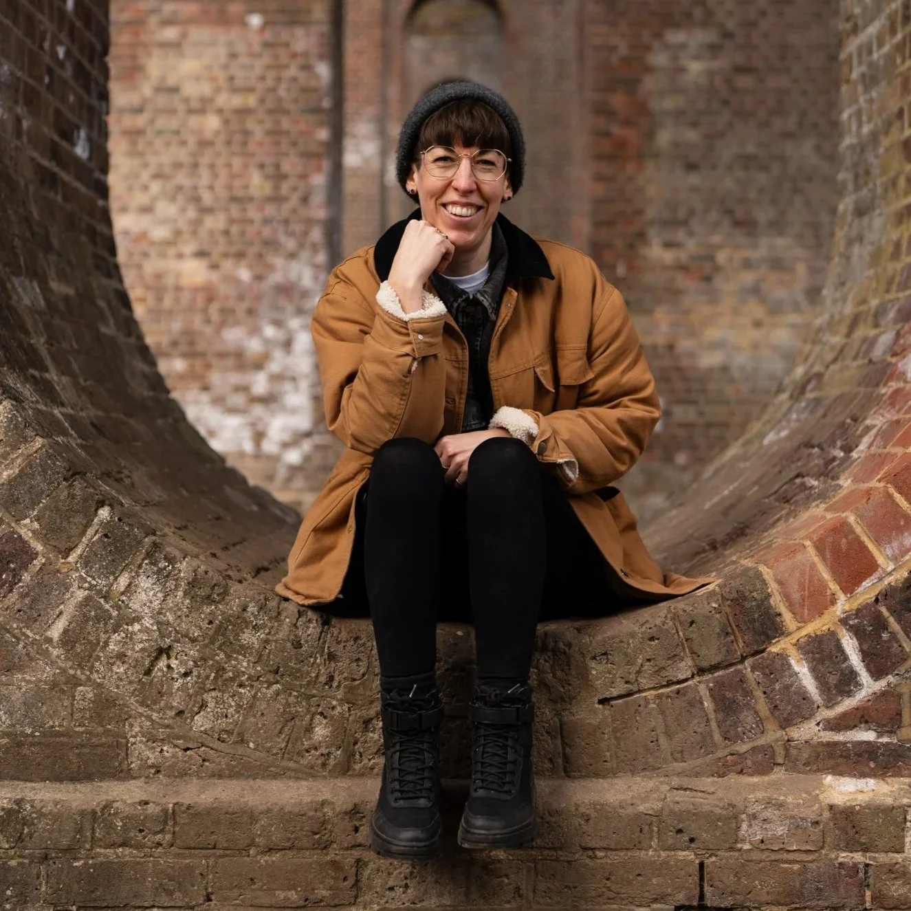 Woman sitting on a curved brick structure with a brick wall background, wearing glasses, a black beanie, a tan jacket with a black collar, black pants, and black boots, smiling at the camera.