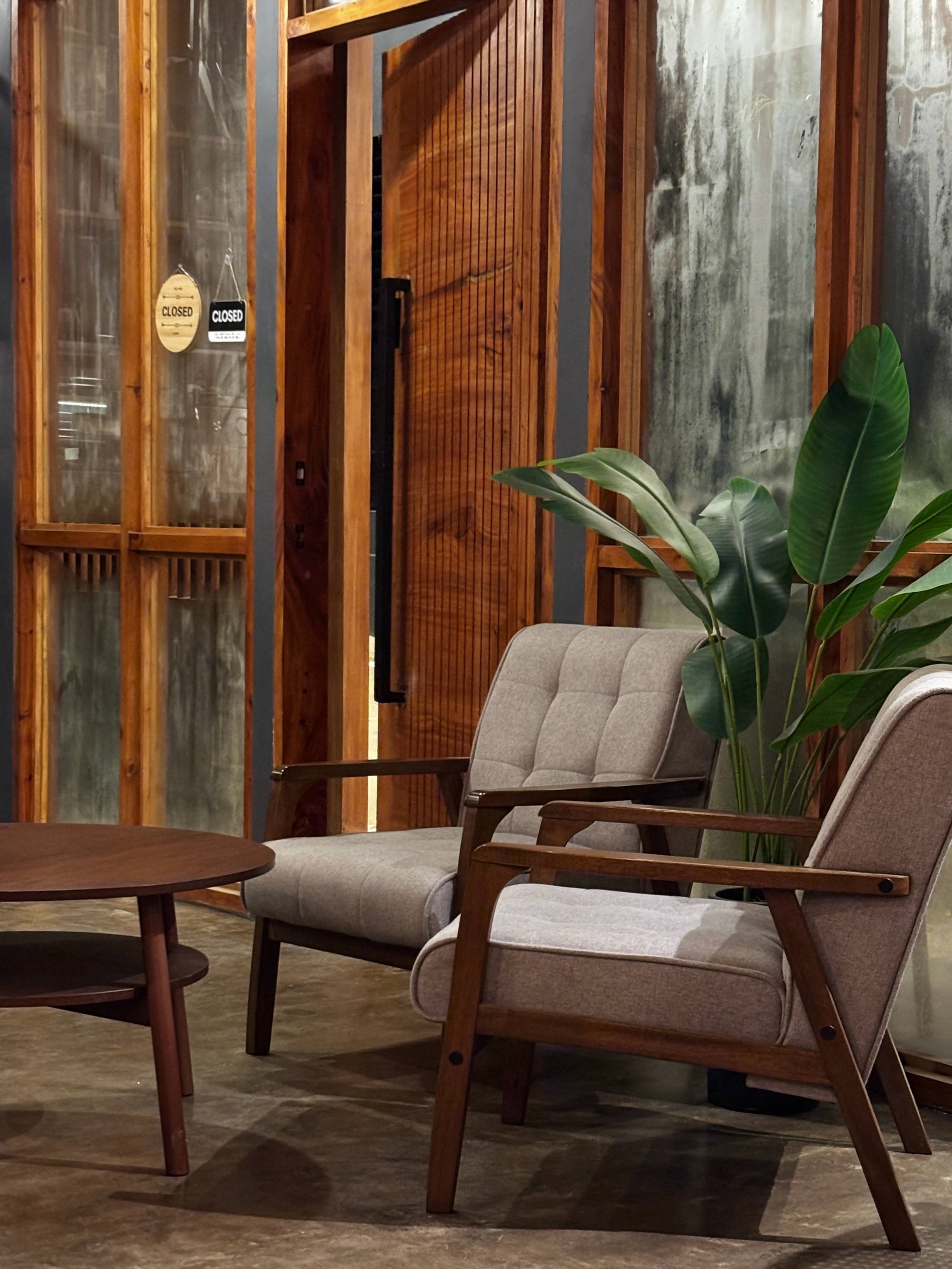 A cozy indoor seating area with a mid-century modern style beige sofa, wooden armchairs, and a round wooden coffee table. A large green leafy plant is beside the sofa, and a textured wall with abstract art is visible in the background. Part of a wooden structure with glass panels and a door is also seen.