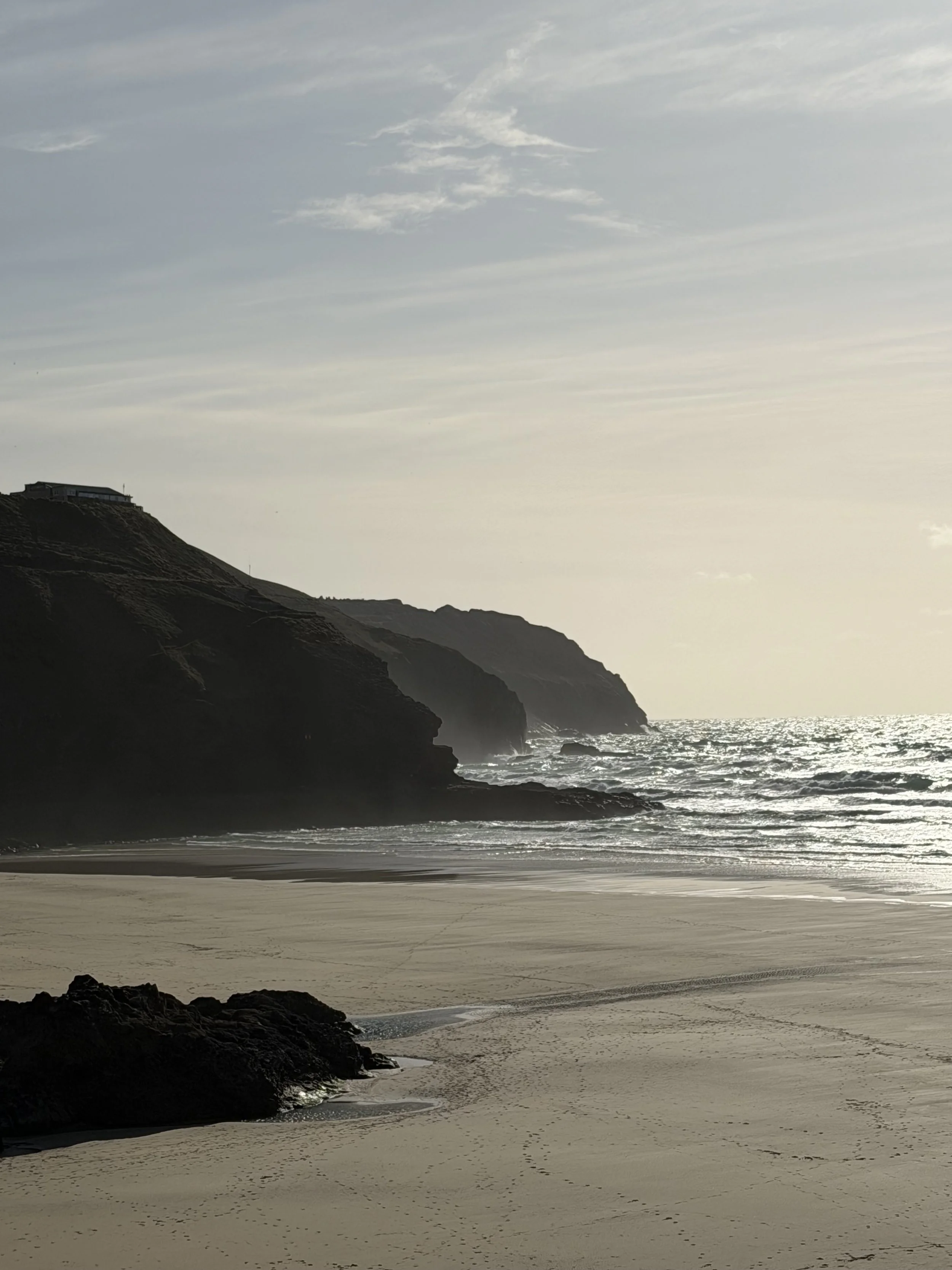 A coastal landscape with cliffs, sandy beach, and ocean waves under a cloudy sky at sunset.