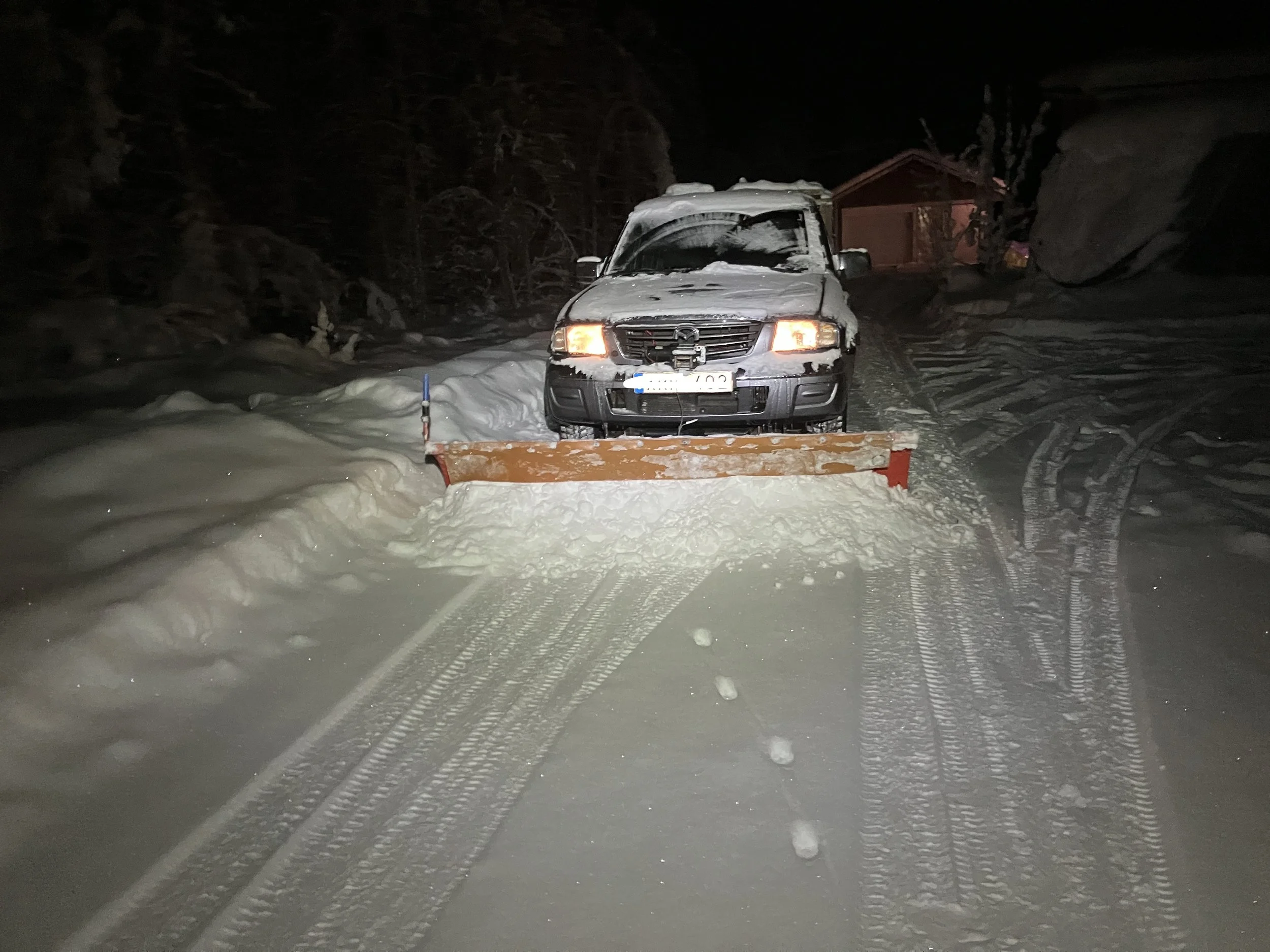 En bil med snö på taket och fronten parkerad framför en snöskopa på en snöig gata om natten. Träd och ett hus är i bakgrunden.