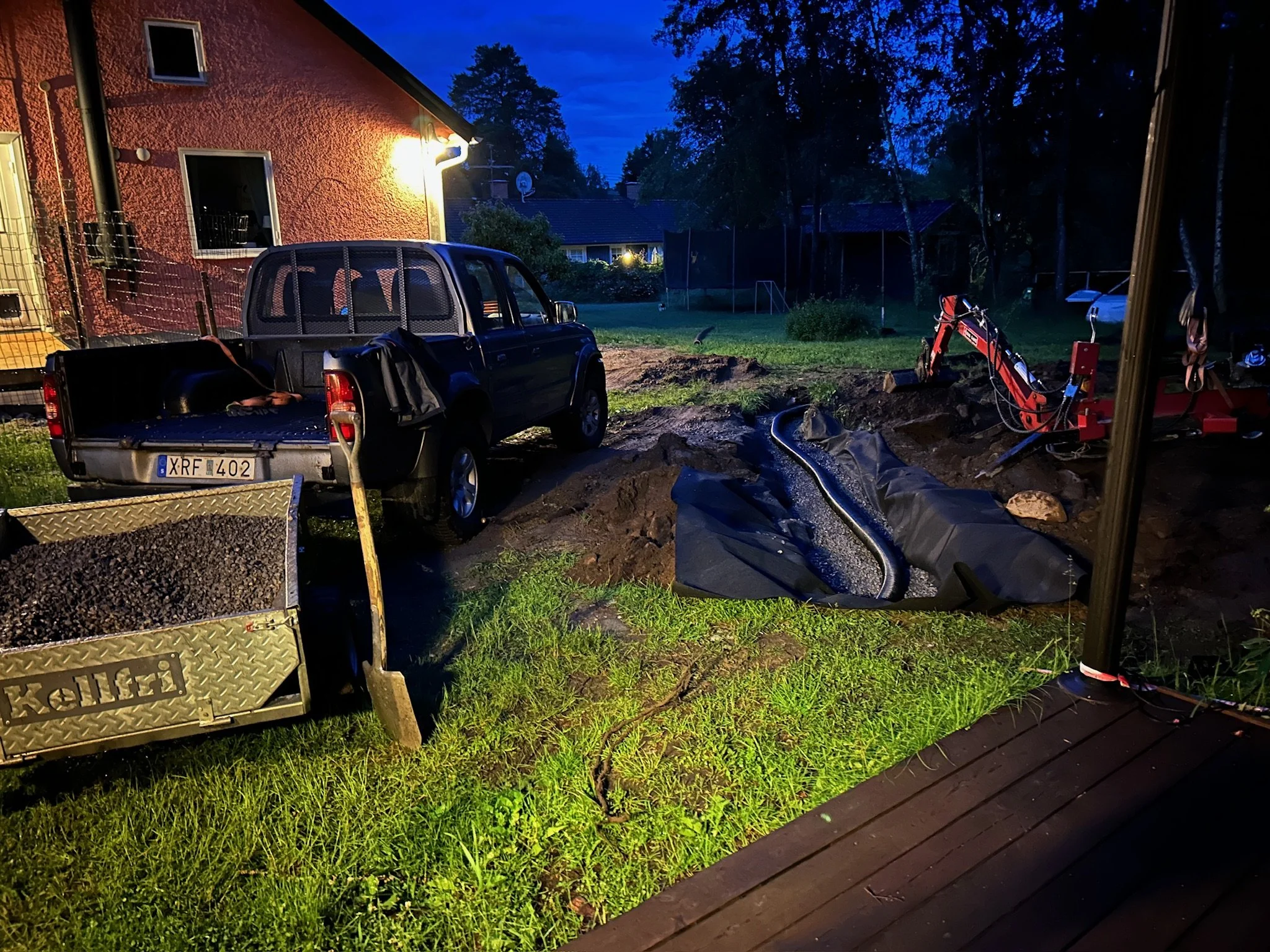 En trädgård på kvällen med en svart pickup, en grävskopa, en trädgårdsslang och ett svart vattenfilter. Det är brunt jord och grönt gräs.