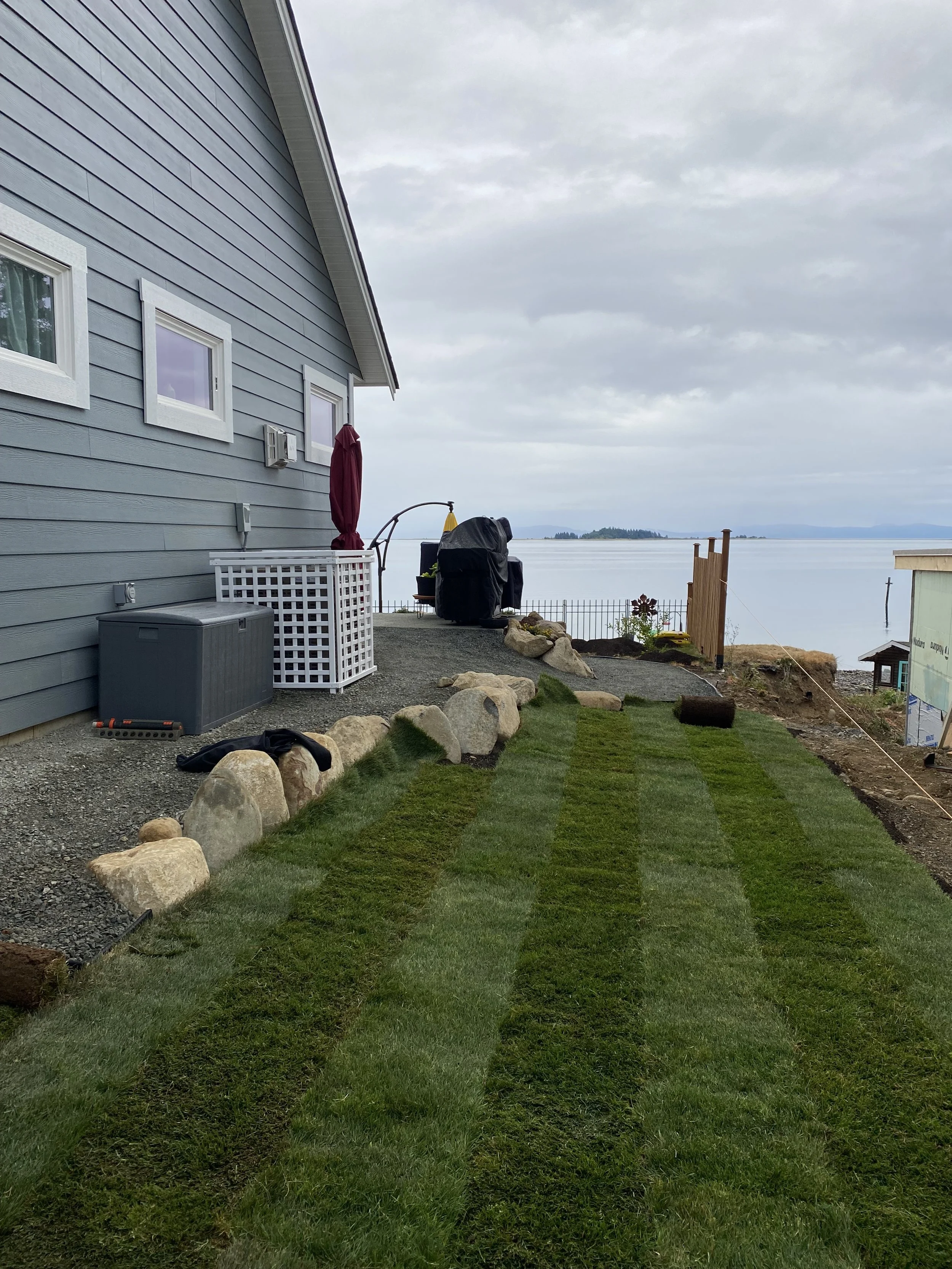 Backyard lawn with freshly laid grass, large rocks along the edge, house with blue siding, and a view of the water and distant land in the background.