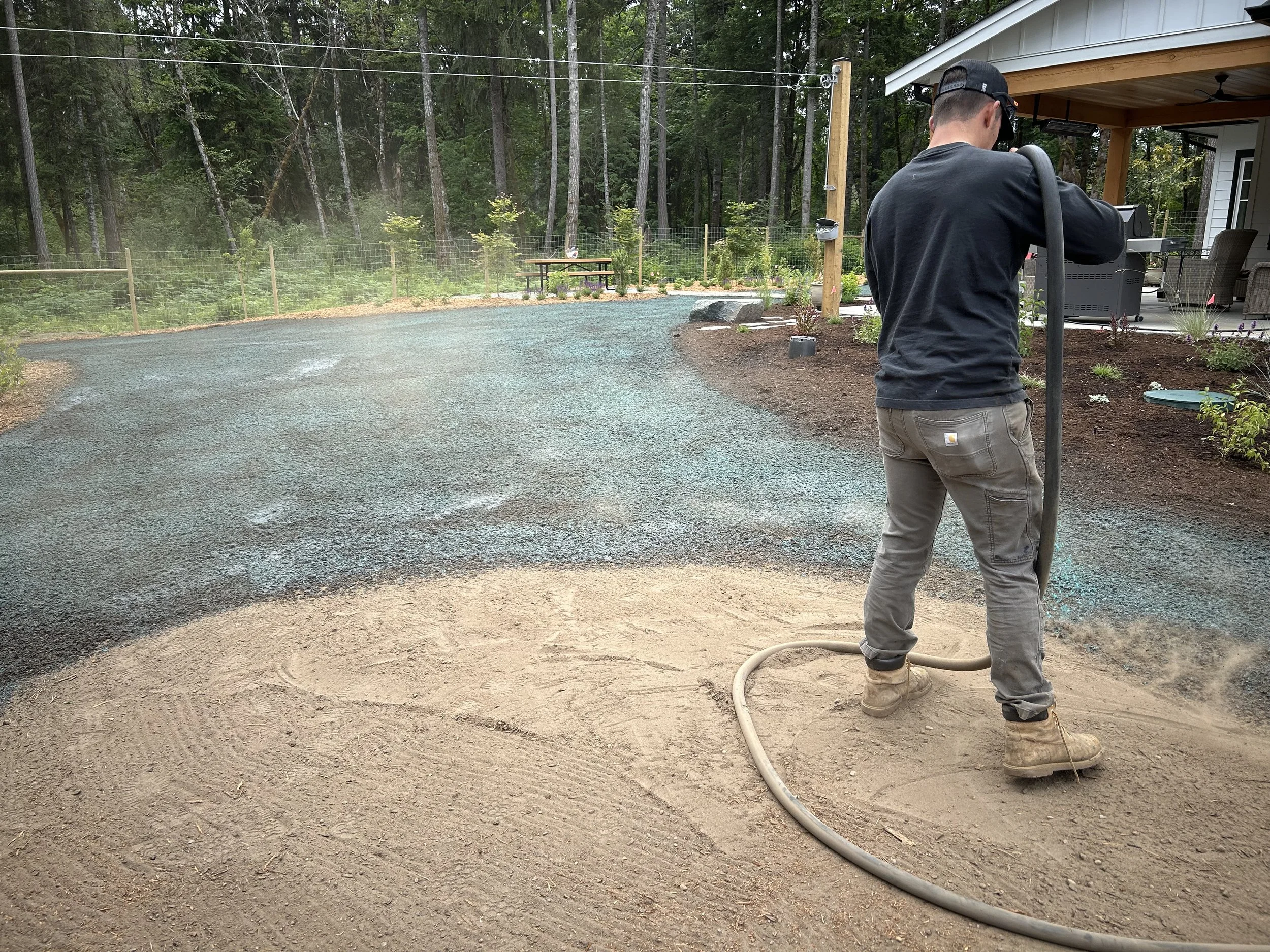 Man operating a vibratory plate compactor on a dirt surface in a backyard, preparing it for landscaping or paving.
