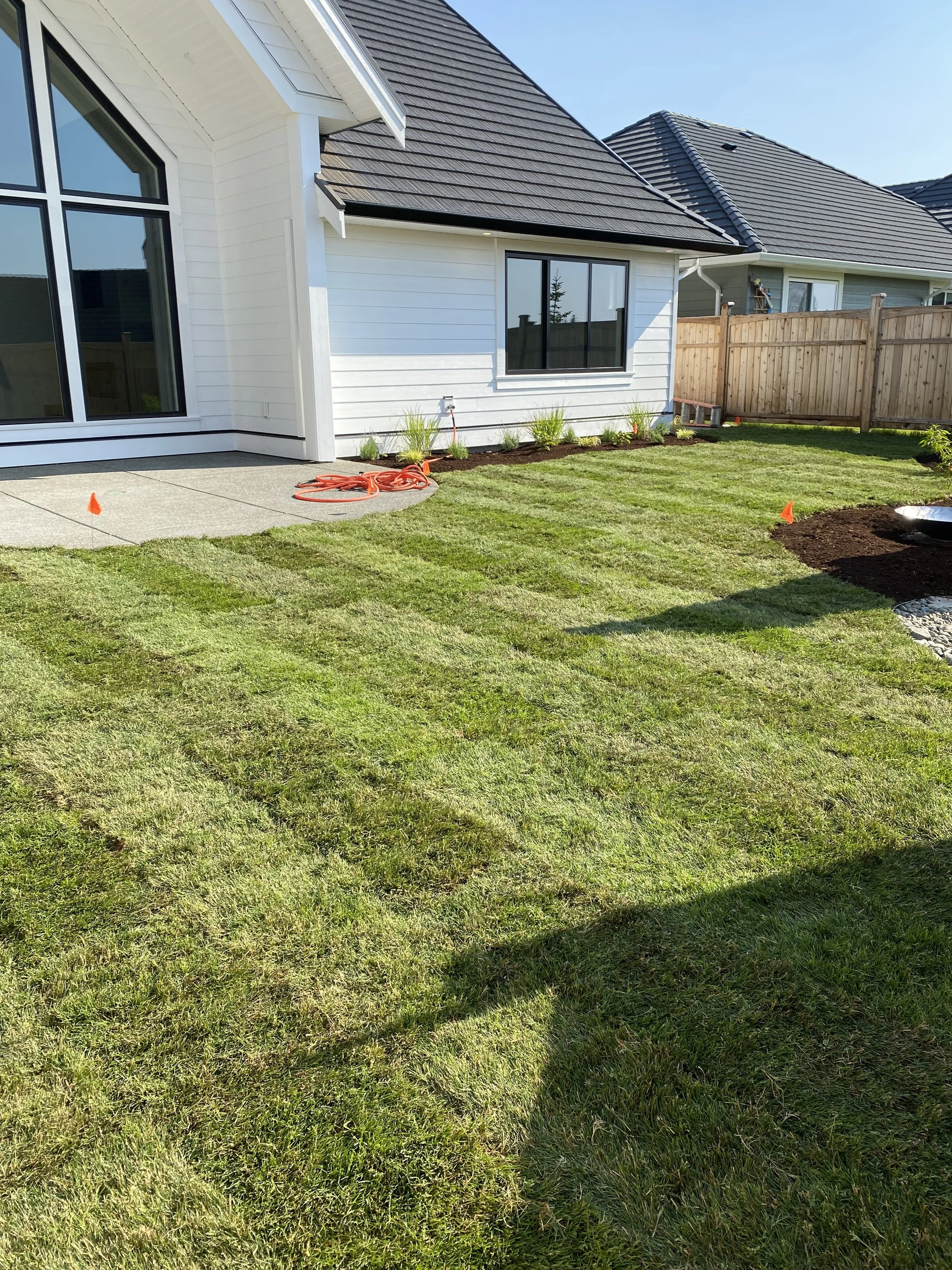 A backyard with freshly laid grass, a concrete patio, and garden bed next to a white house with large windows and a wooden fence.