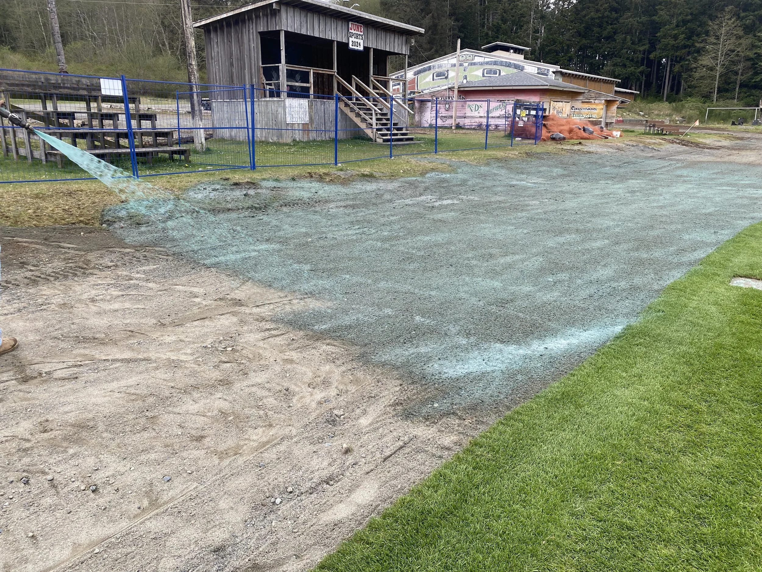 A construction or landscaping project in progress, with a section of ground being graded or prepared, a hose spraying water, a wooden building with stairs and a sign reading 'June Sports 2024', and a fenced area with a couple of structures and trees 