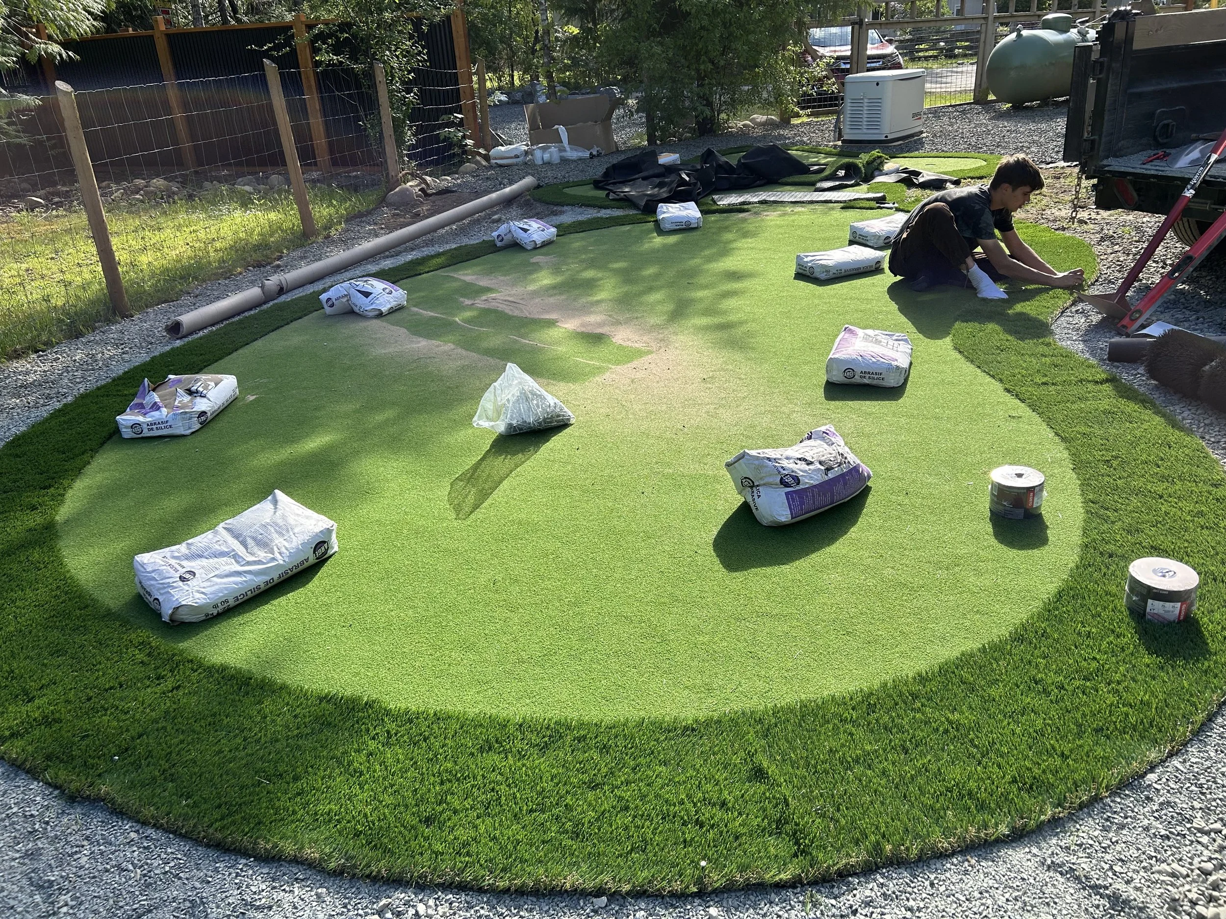 Person installing artificial grass on a backyard lawn, with bags of materials and tools around.