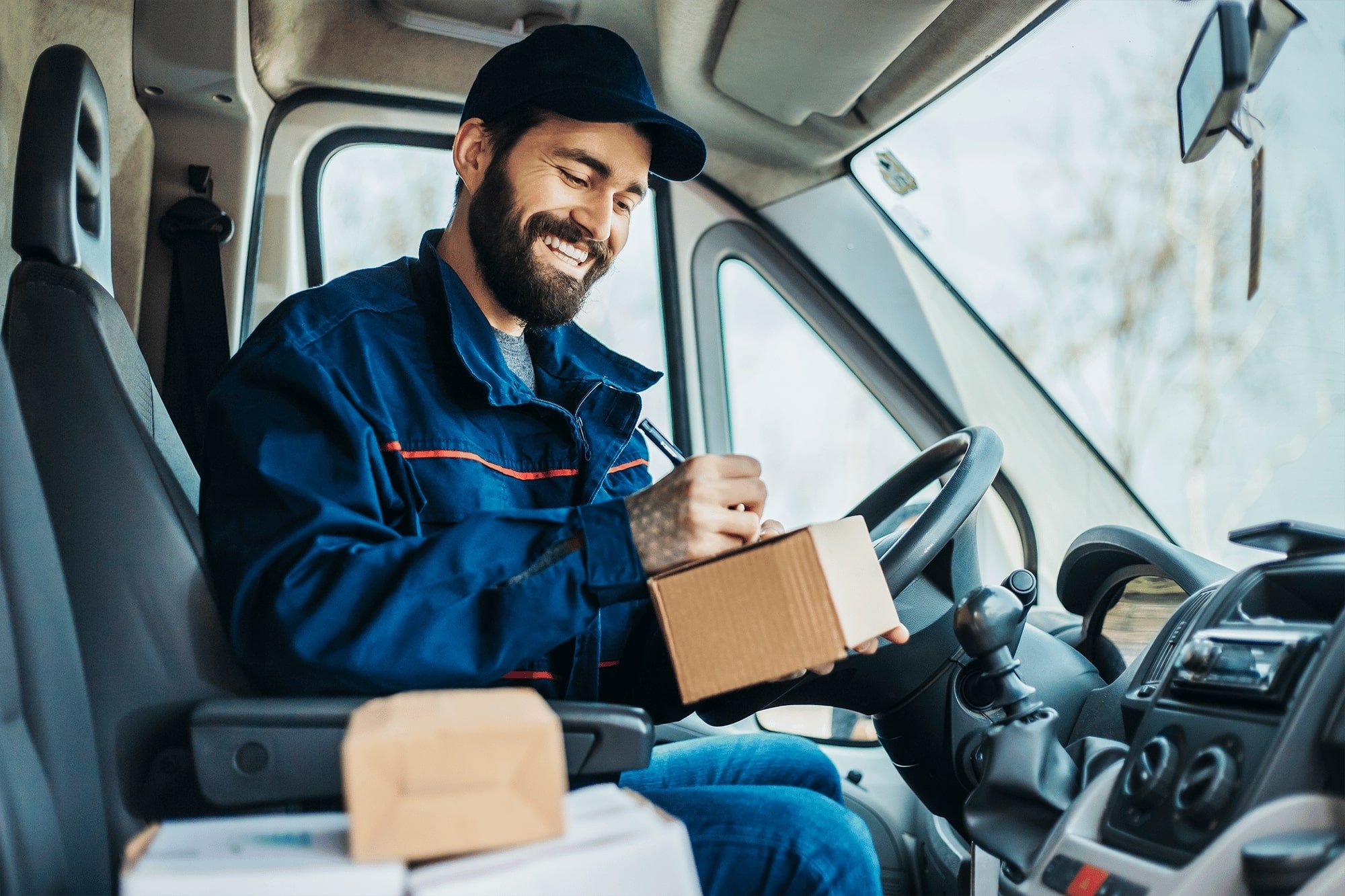 A smiling delivery driver with a beard and a hat, sitting in the driver's seat of a delivery truck, holding and signing for a package.