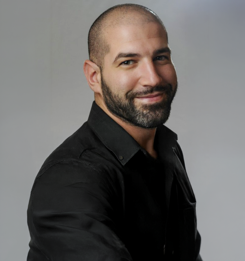 Portrait of a smiling man with a beard and a shaved head, wearing a black shirt against a light gray background.