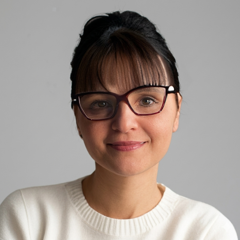 A woman with dark hair, wearing glasses with a dark frame and a white sweater, smiling at the camera against a plain background.
