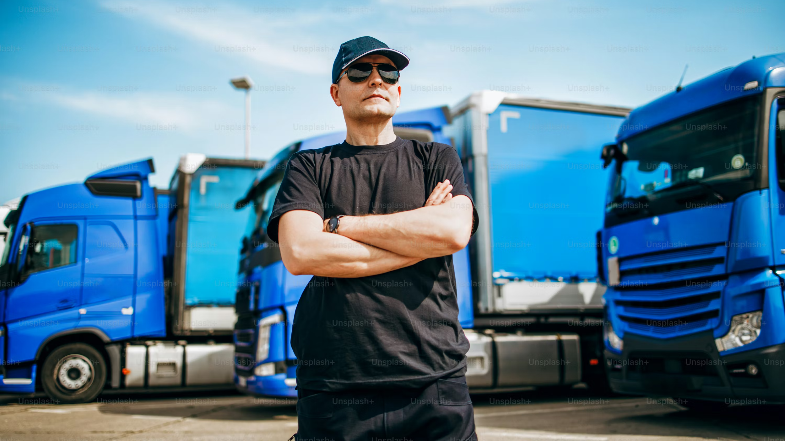 A man wearing sunglasses, a black t-shirt, and a cap stands with arms crossed in front of a row of blue trucks on a sunny day.