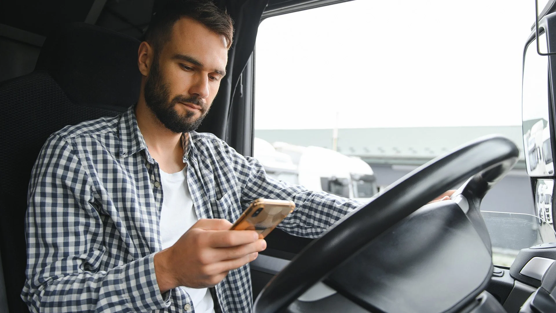 Man with beard wearing checkered shirt sitting in driver's seat of truck, using smartphone.