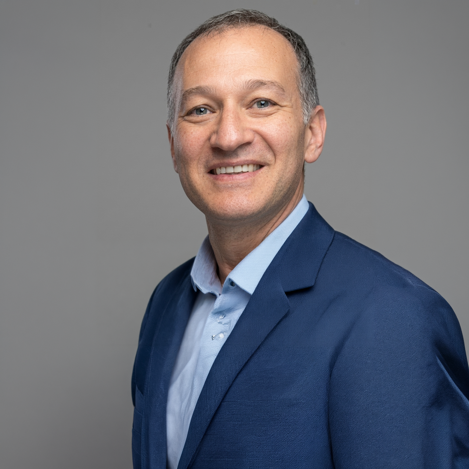 Headshot of a smiling man in a blue suit and light blue shirt against a gray background.