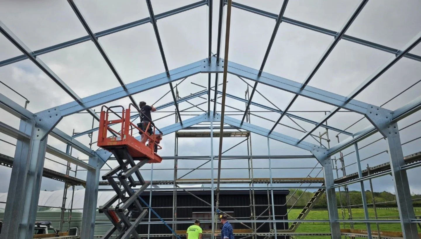 Construction workers building a metal framework, with one worker operating a scissor lift and two workers on the ground discussing near a partially assembled structure outdoors with a grassy field and cloudy sky in the background.