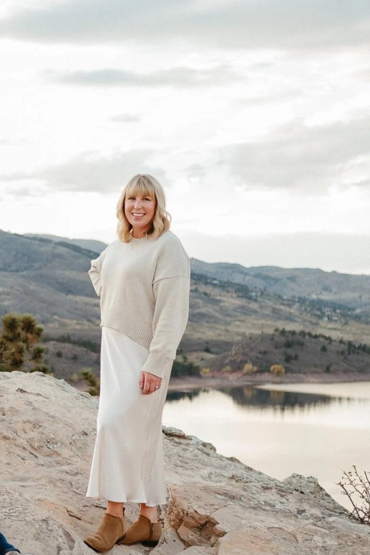 A woman standing on a rocky shore near a lake with mountains in the background, wearing a beige sweater, white dress, and tan ankle boots, smiling at the camera.