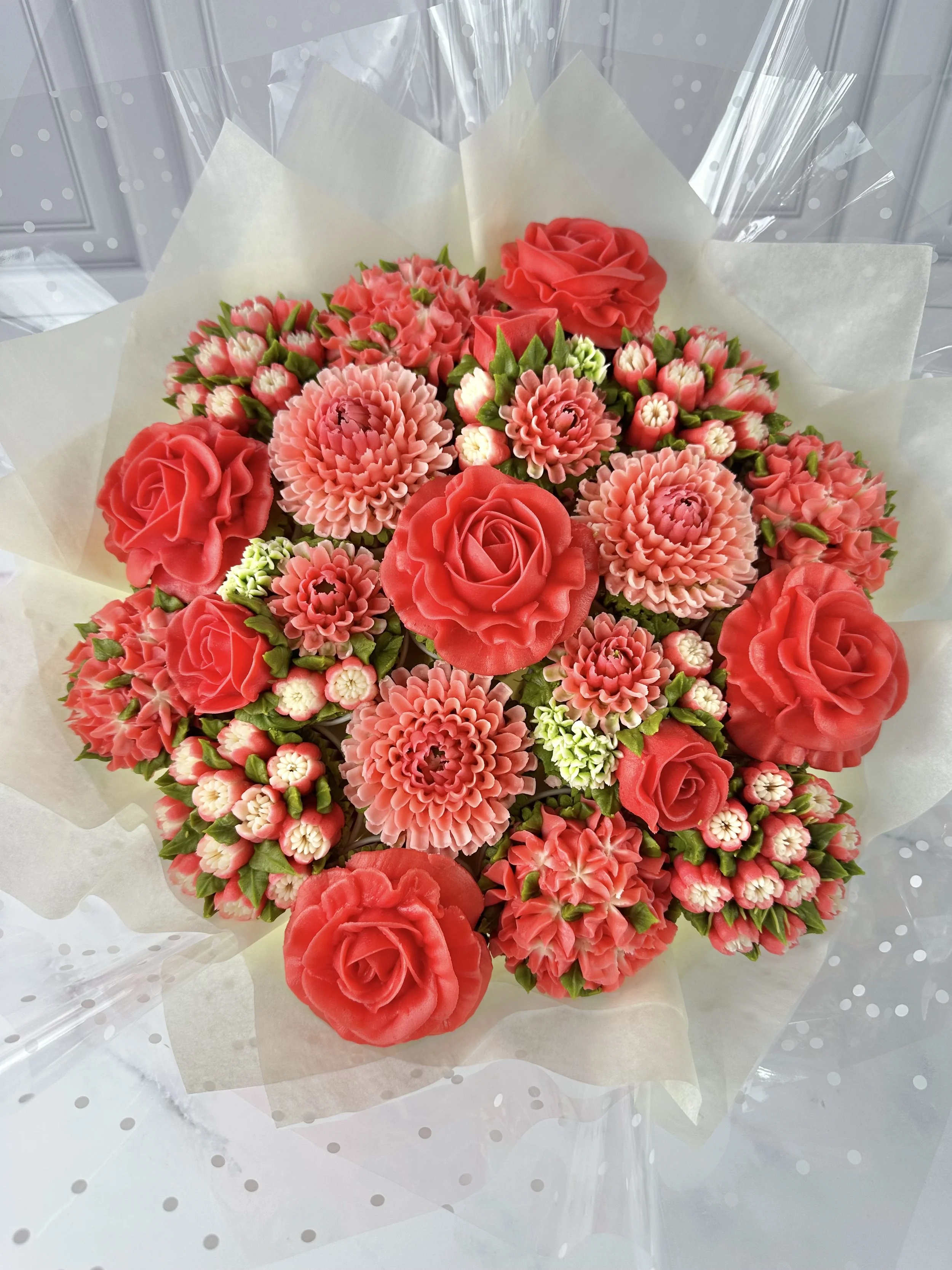 A bouquet of pink and red flowers, including roses and carnations, wrapped in clear plastic and white tissue paper.