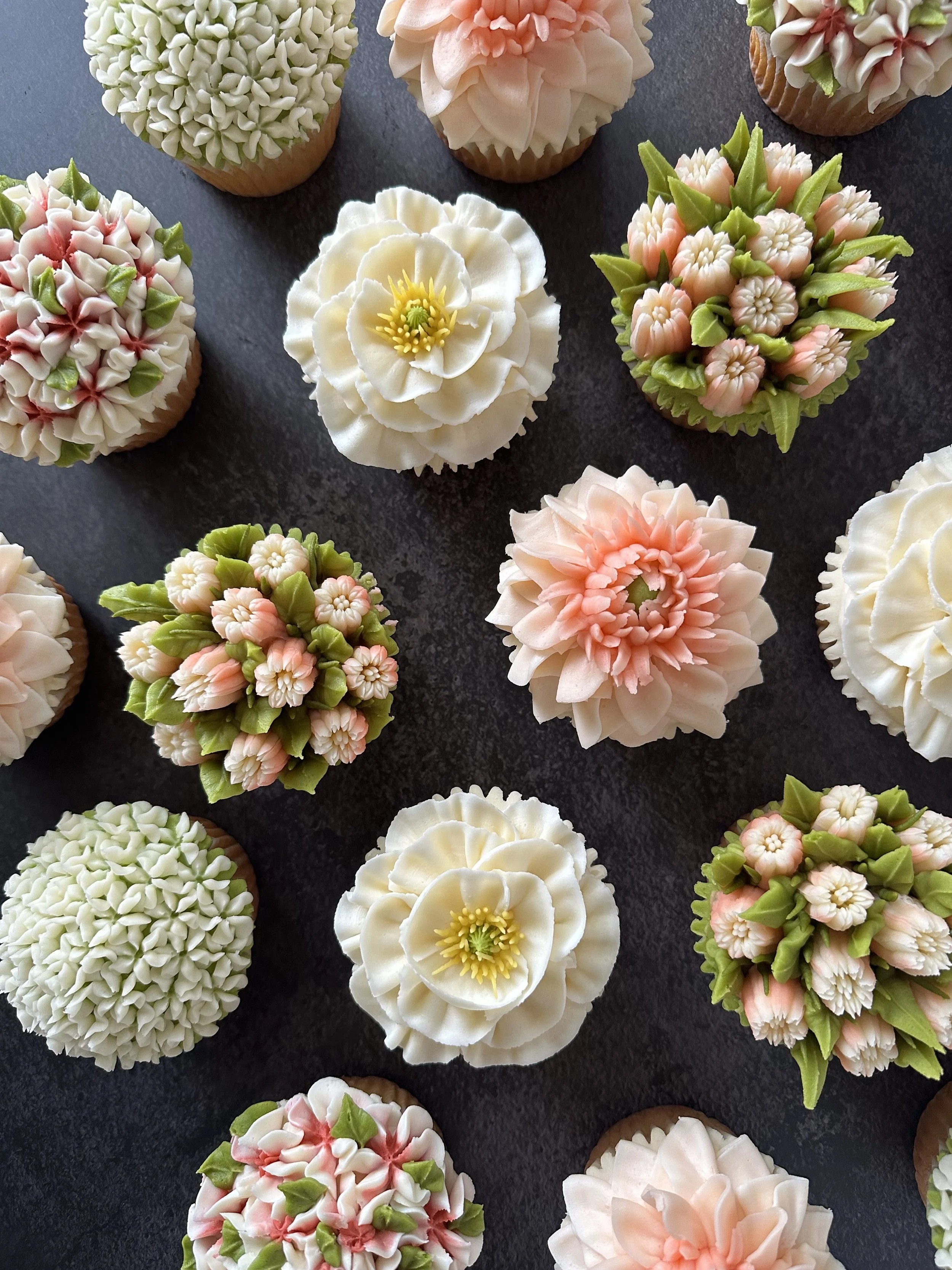 Assorted decorated cupcakes with floral and leafy frosting on a dark surface.