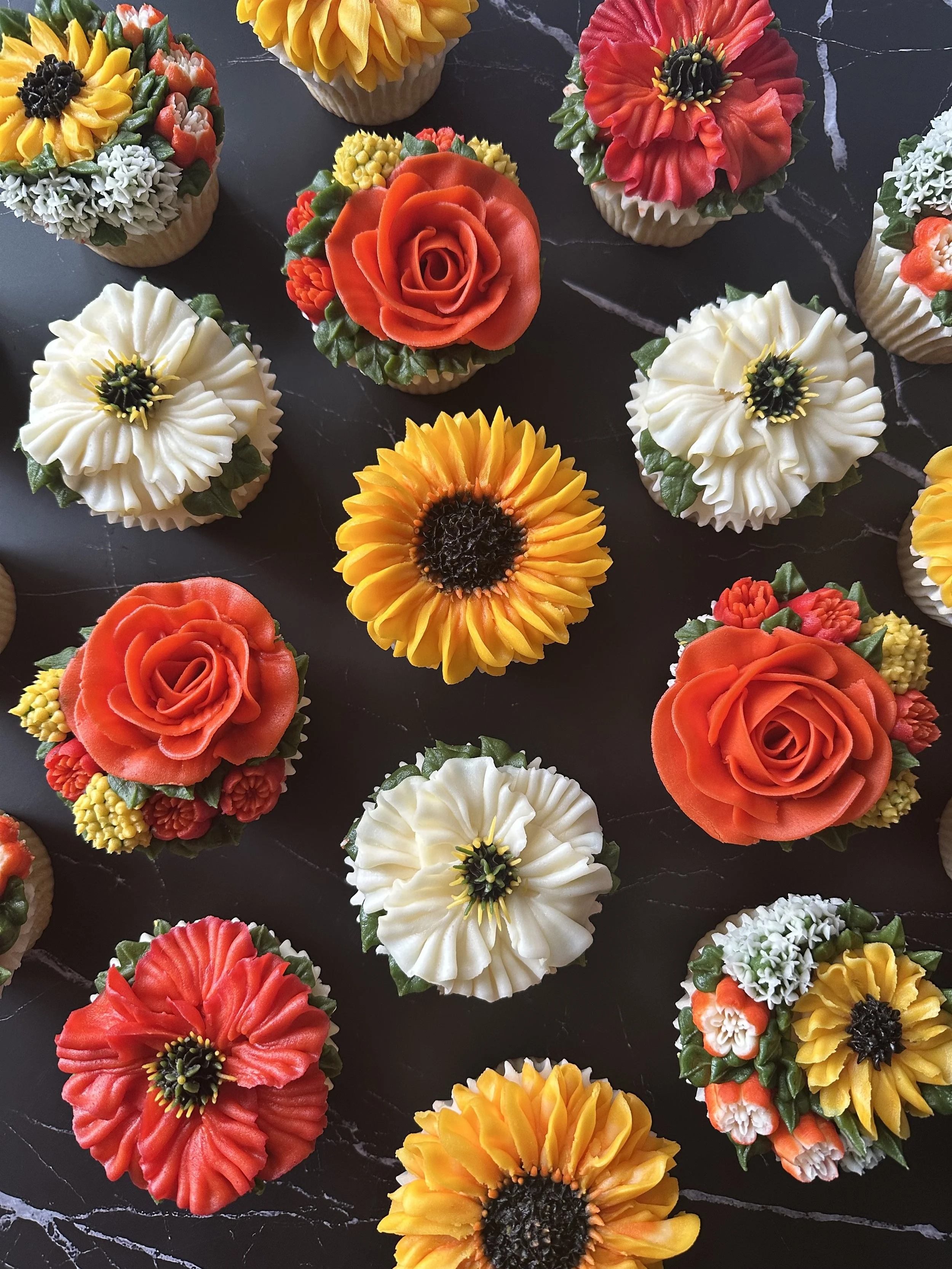 Assorted cupcakes decorated with flowers, including sunflowers, roses, poppies, and daisies, on a dark marble surface.