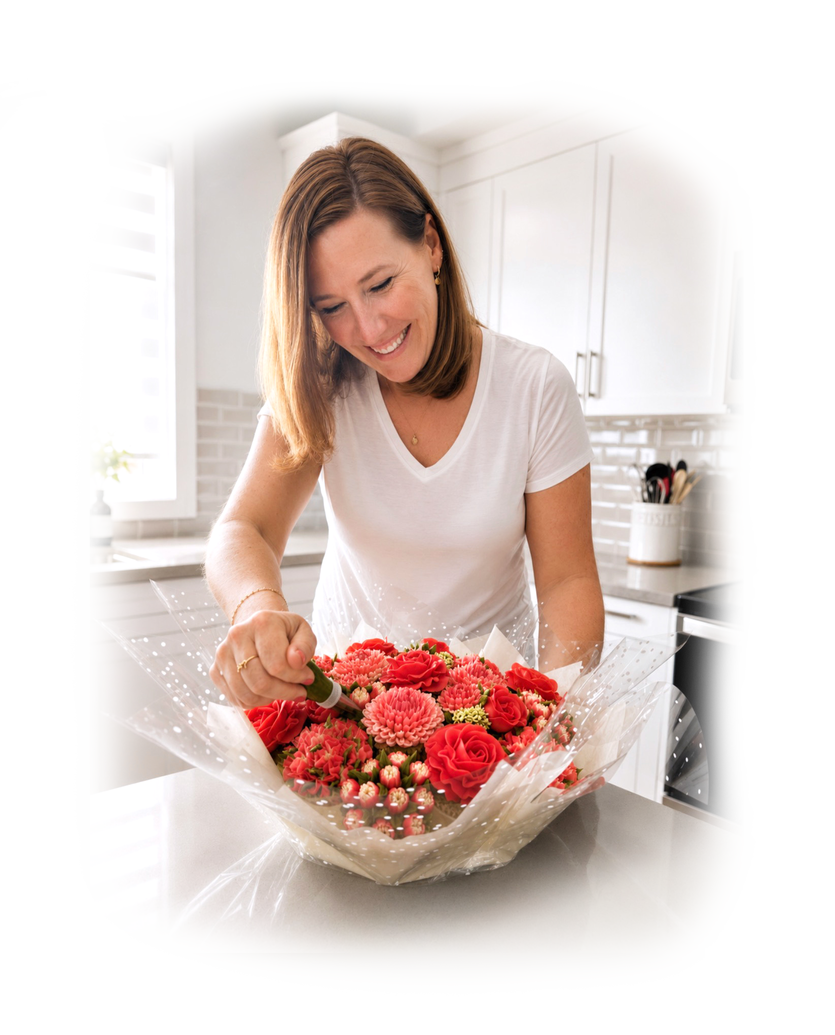 A woman in a white shirt smiling as she arranges a bouquet of bright pink and red flowers in a kitchen.