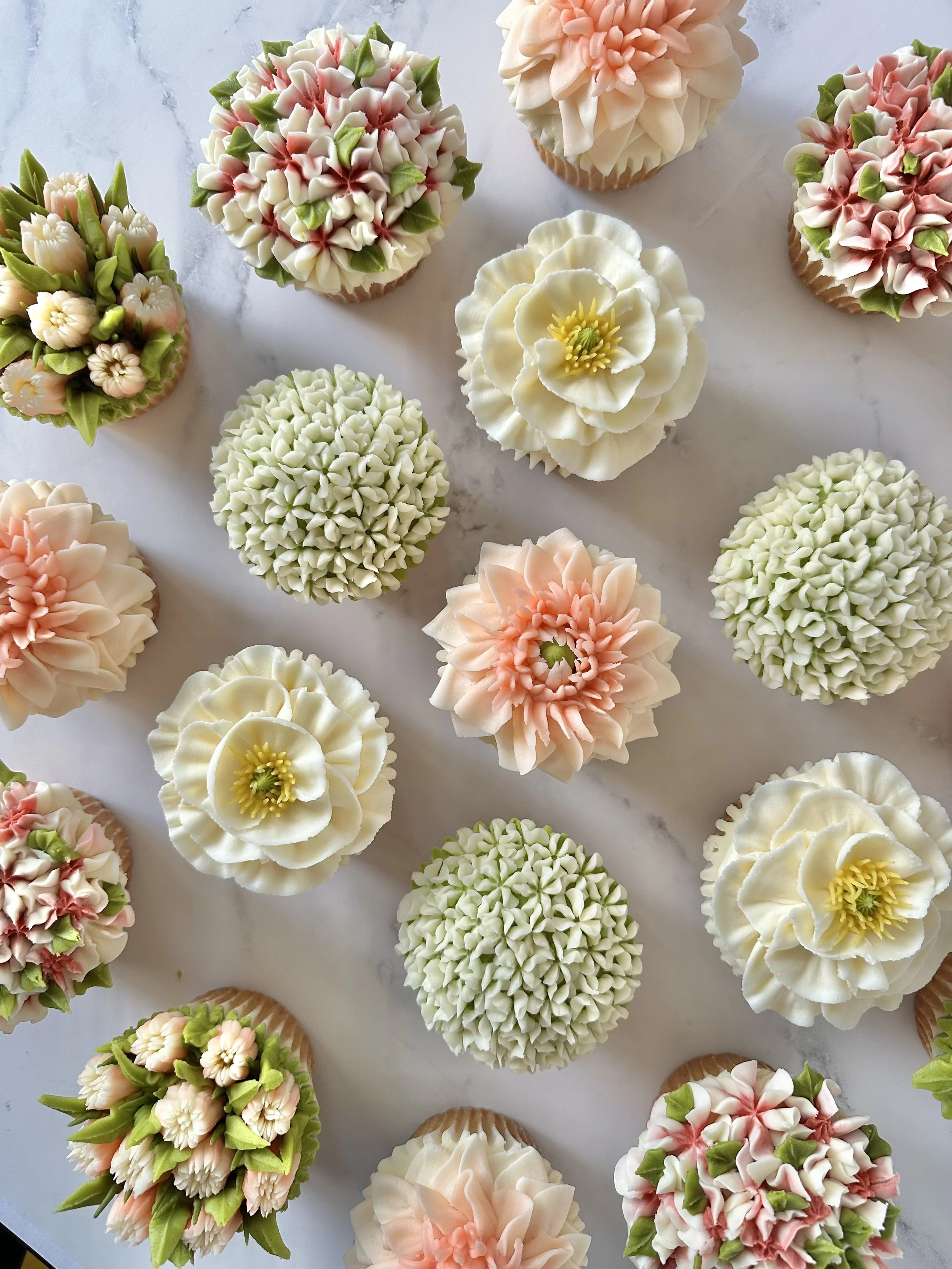 Assorted cupcakes decorated with intricate floral frosting designs in pastel colors, arranged on a light marble surface.