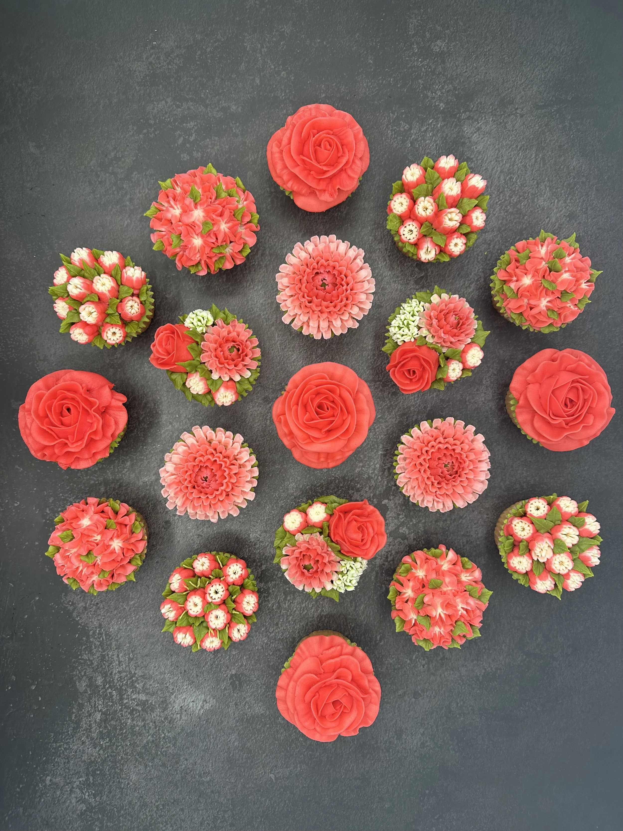 Assorted floral cupcakes with pink, red, and white icing decorated to resemble roses and other flowers, arranged on a dark textured surface.