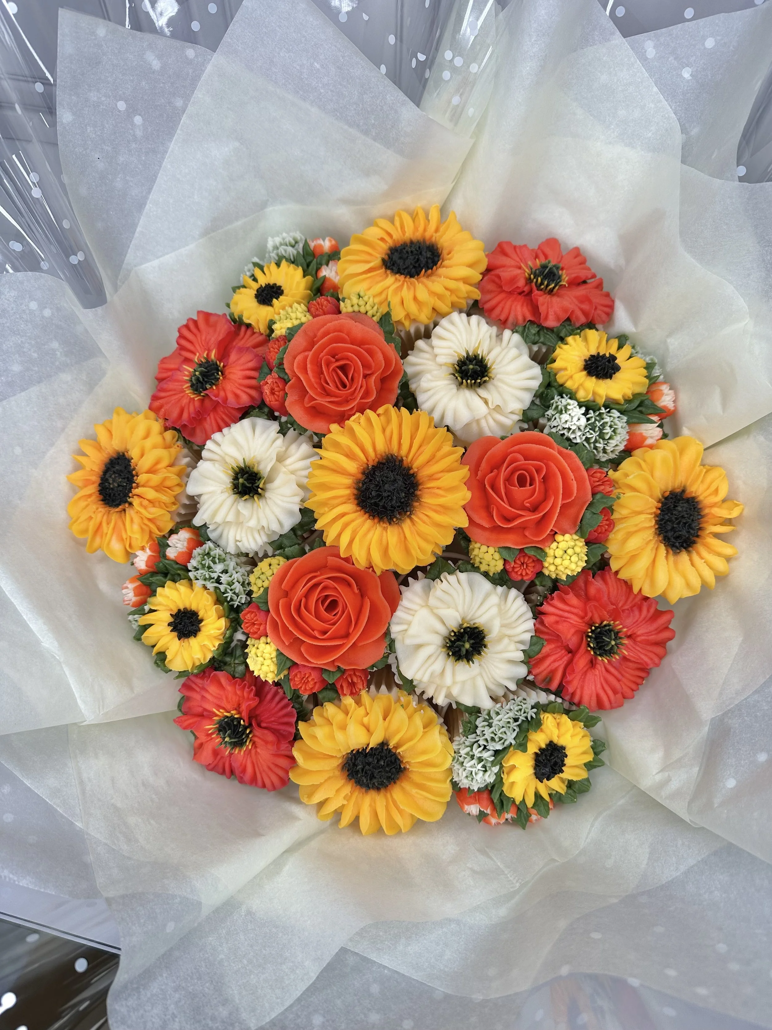A bouquet of colorful artificial flowers, including orange roses, yellow sunflowers, white daisies, and small clusters of orange and white flowers, wrapped in white tissue paper.