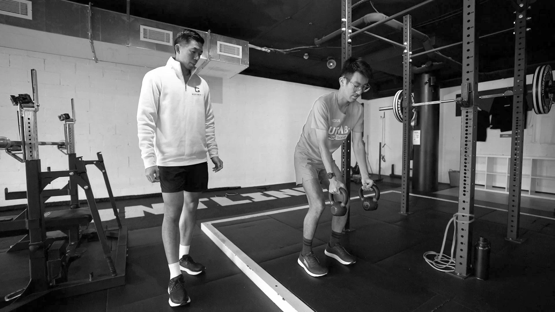 A man in athletic clothes is doing a kettlebell exercise in a gym while another man gawking.