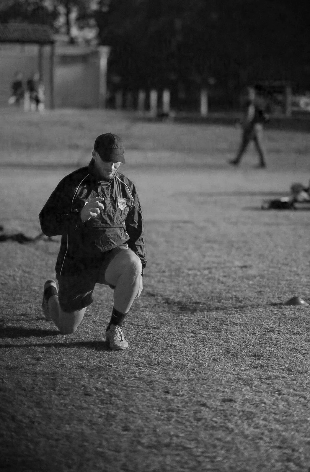 A man kneeling on one knee on a sports field, lacing his shoe, wearing a sports jacket, shorts, and a cap, in a black-and-white photo taken during the evening or night.