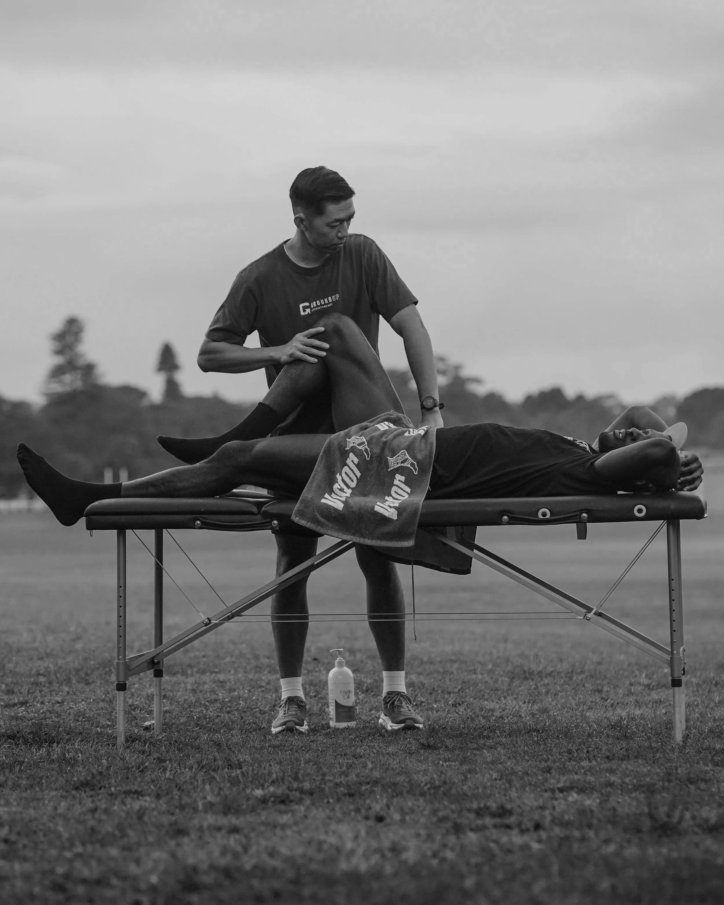 A person receiving a leg massage on a portable massage table outdoors, with a towel covering part of their body, and an individual providing the massage. The scene is set in a grassy field during daytime.