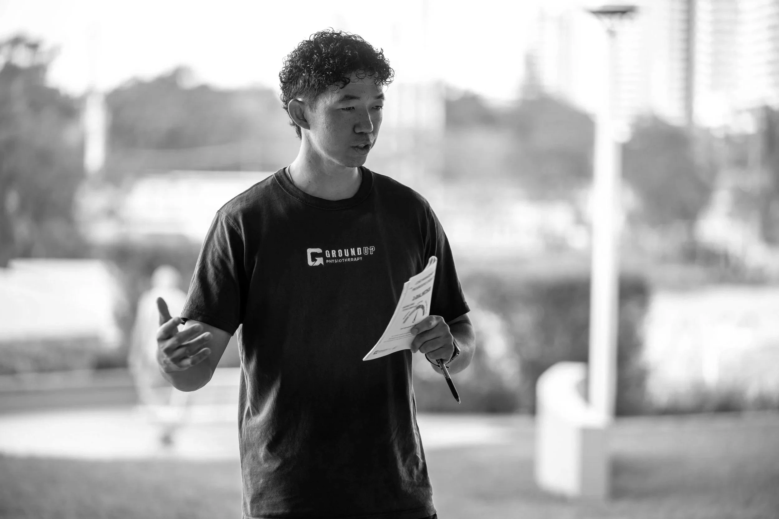 A young man with curly hair wearing a GroundUp Physiotherapy T-shirt, holding a pamphlet and speaking outdoors with a blurred cityscape background.