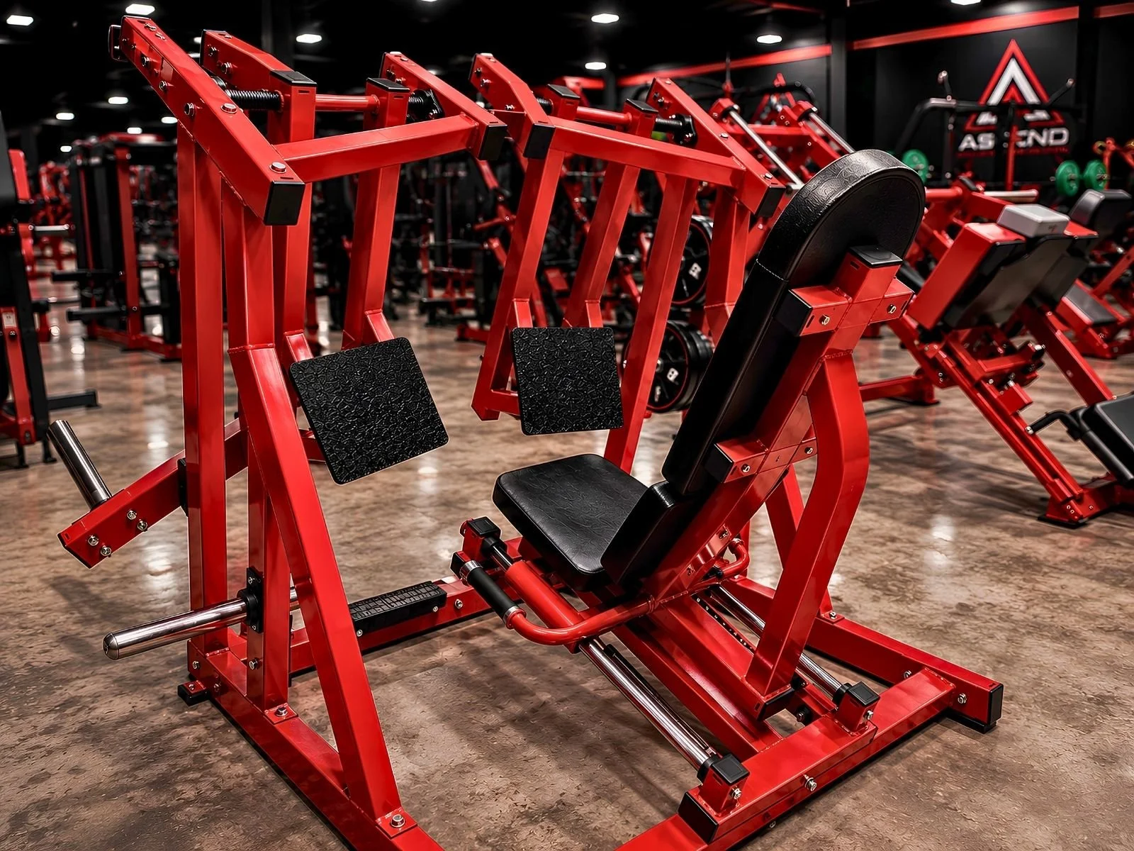 Red strength training machine with black padded seat and foot pedals in a gym with wooden flooring.