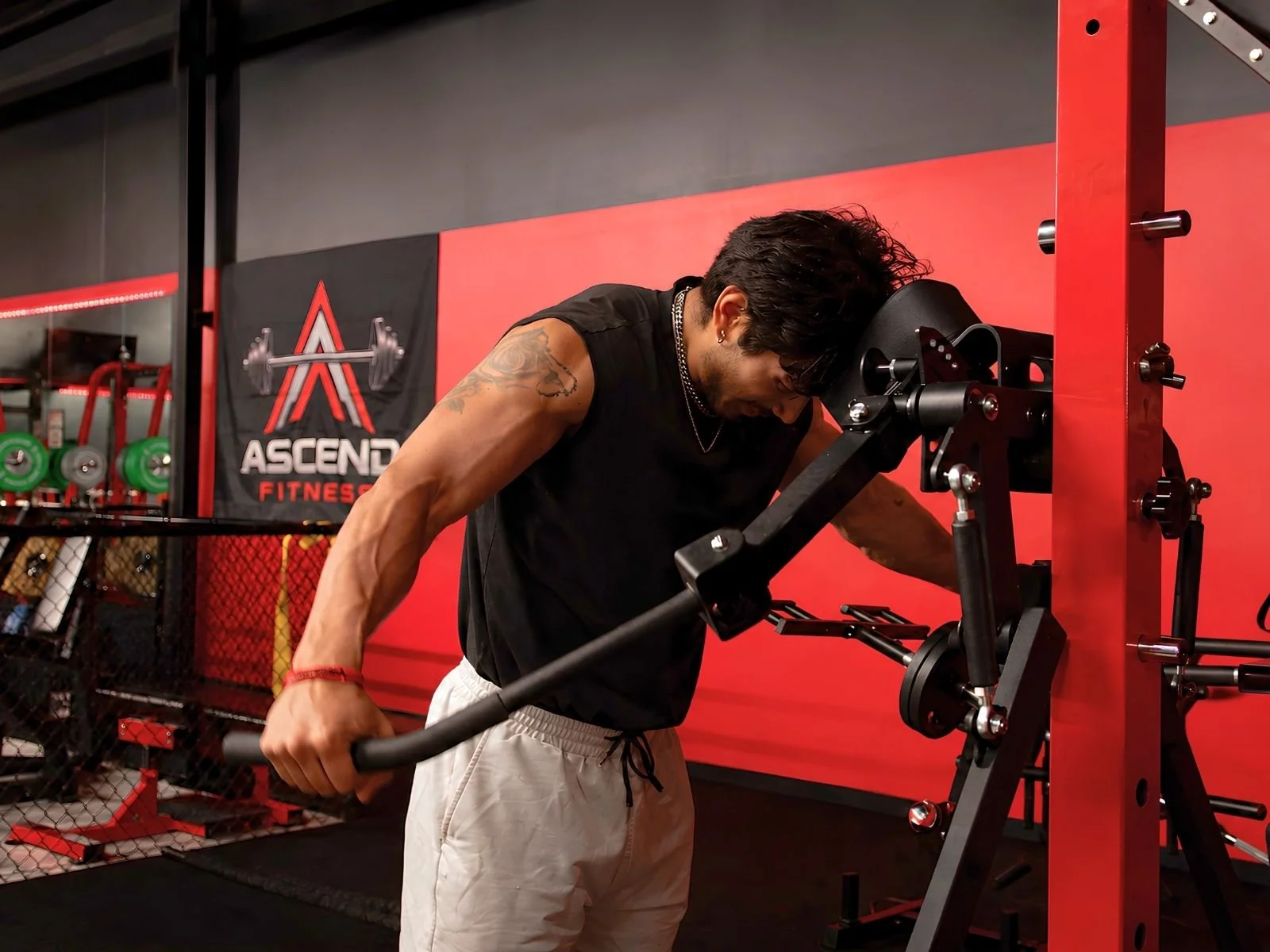 A man with tattoos and a red bracelet is performing a workout at a gym, leaning on a piece of exercise equipment. The gym has a red and black color theme, with fitness branding on the wall in the background.