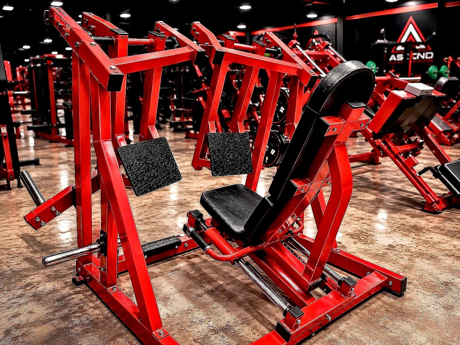 Red strength training machine with black padded seat and footrests in a gym with various other equipment in the background.