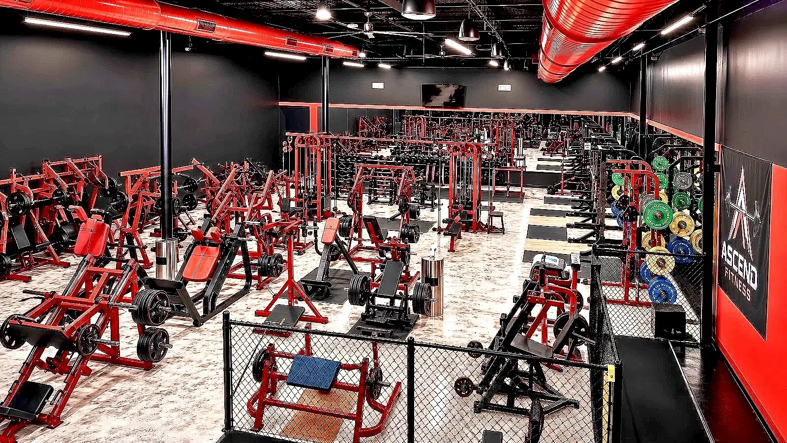 A modern gym filled with exercise equipment, including weight machines, benches, free weights, and weight plates. The gym has a black wall with a large mirror and a wall-mounted TV.
