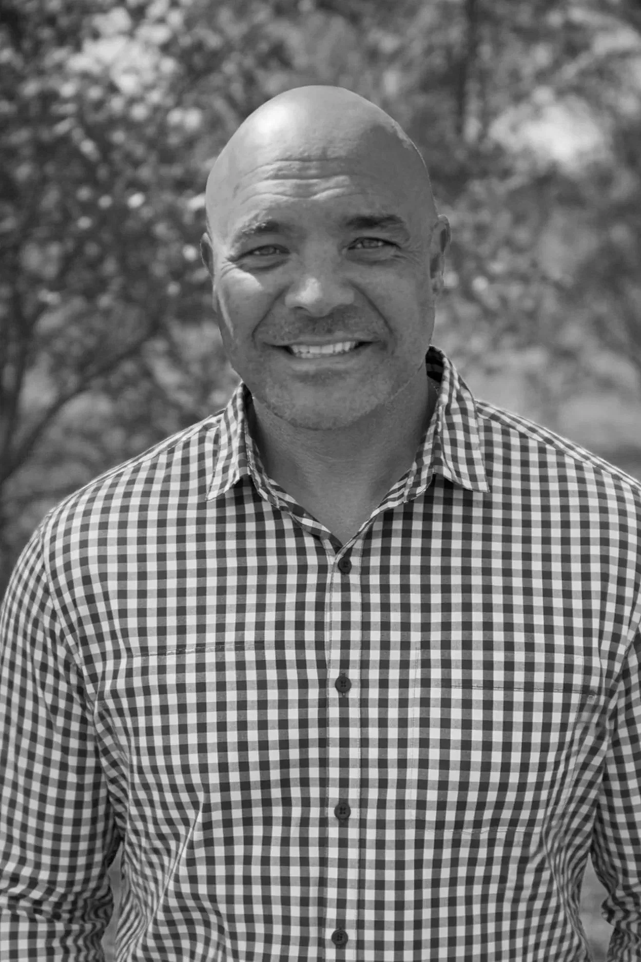 A smiling bald man wearing a checkered button-up shirt outdoors with trees in the background.