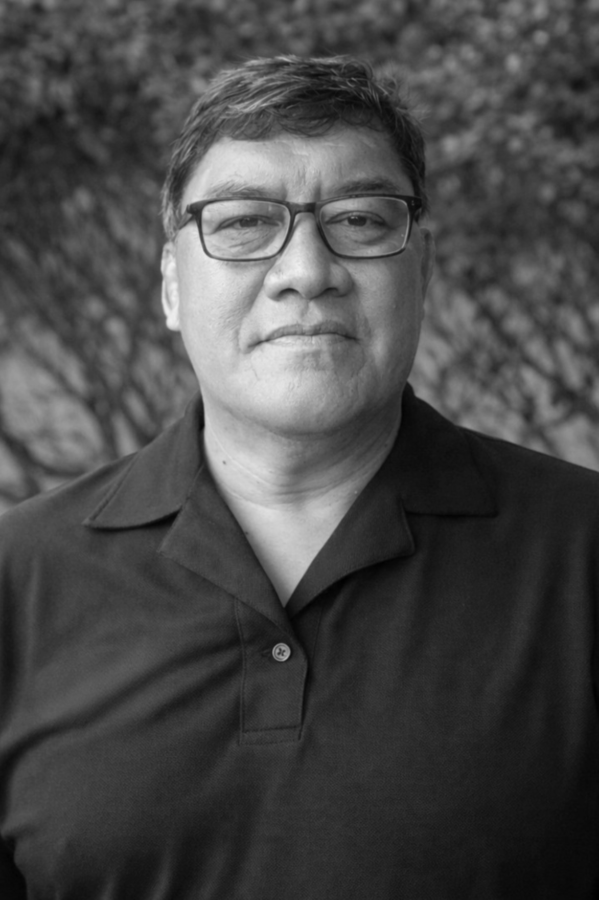 A black-and-white portrait of an Asian man wearing glasses and a polo shirt, standing outdoors in front of blurred foliage.