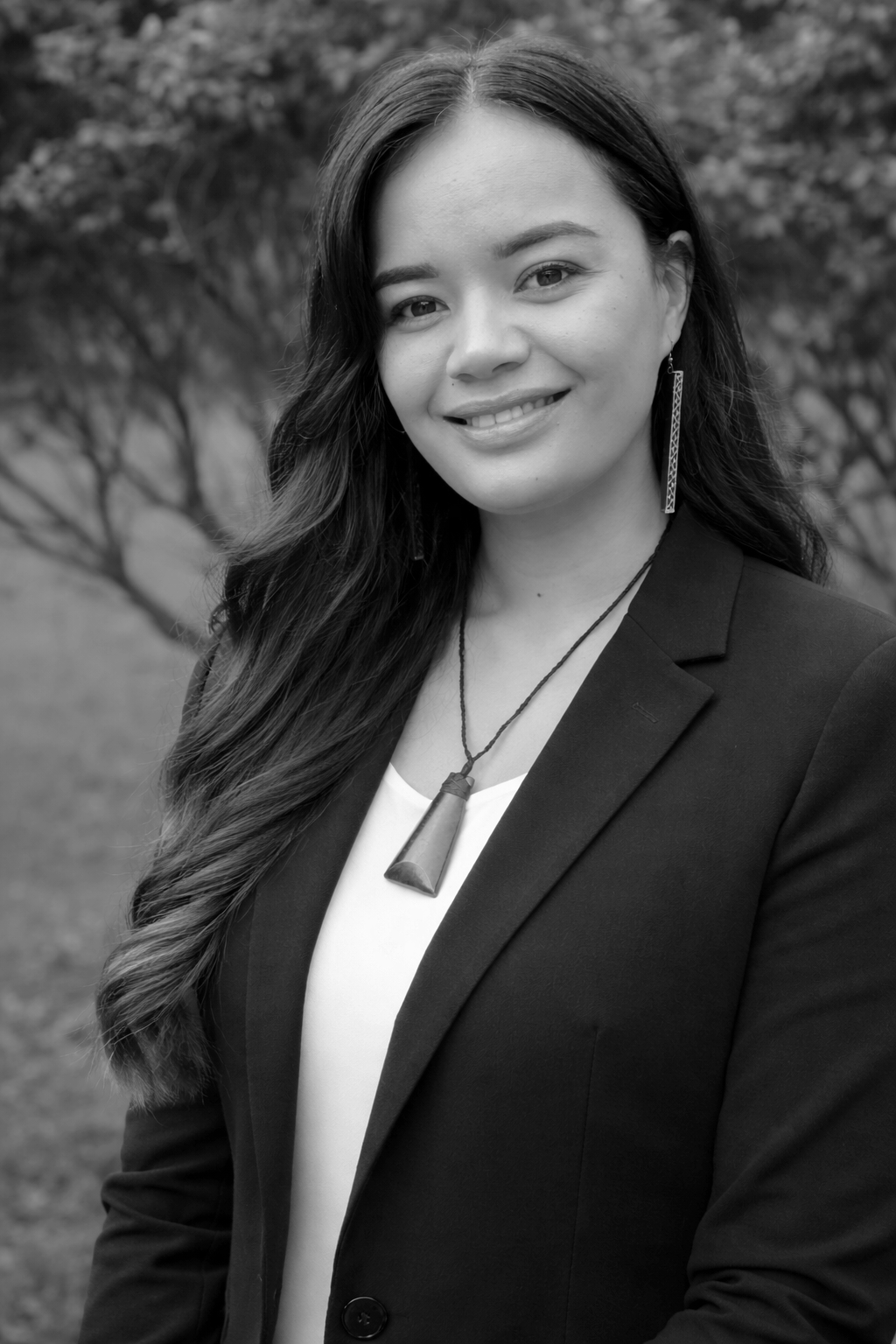 A woman with long hair smiling outdoors, wearing a blazer, white shirt, earrings, and a necklace with a large pendant, background of trees.