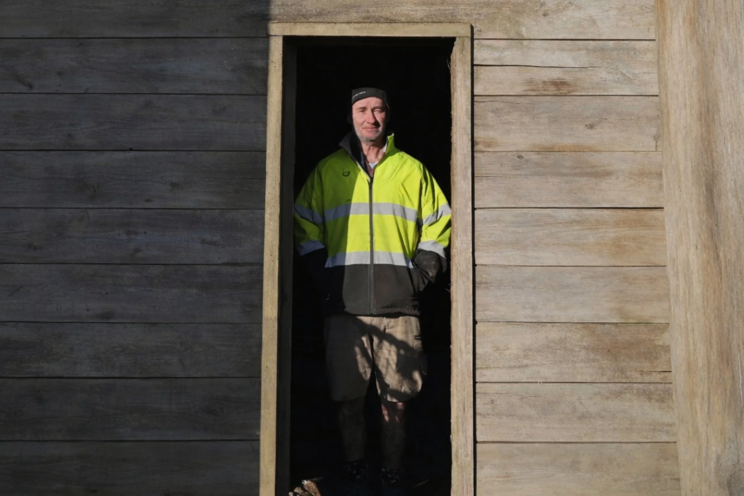A man standing in a doorway between wooden walls, wearing a high-visibility yellow jacket, shorts, and a black beanie.