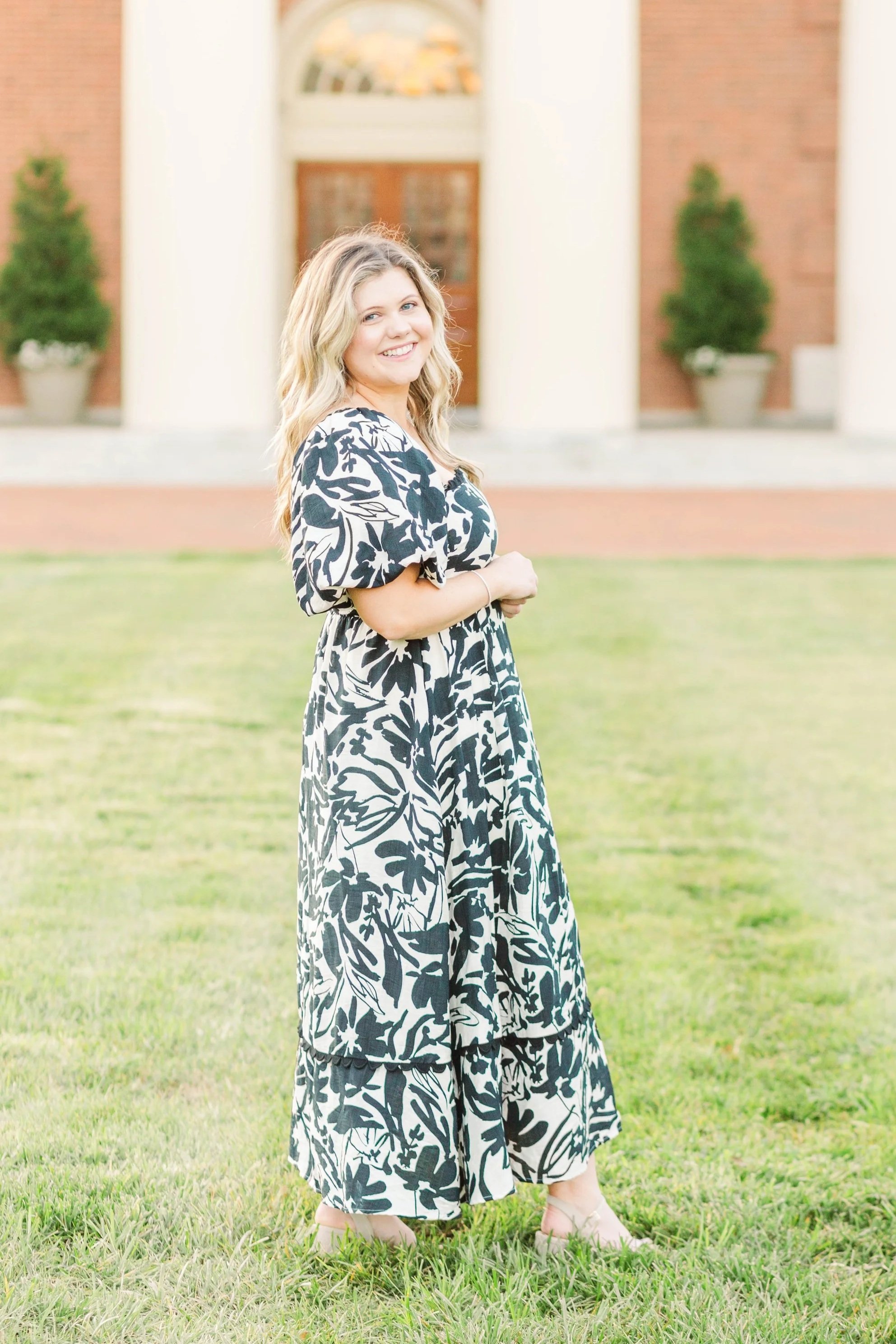 A woman standing outdoors on a grassy area in front of a building with brick and beige columns, wearing a black and white floral dress, smiling, with blonde wavy hair.