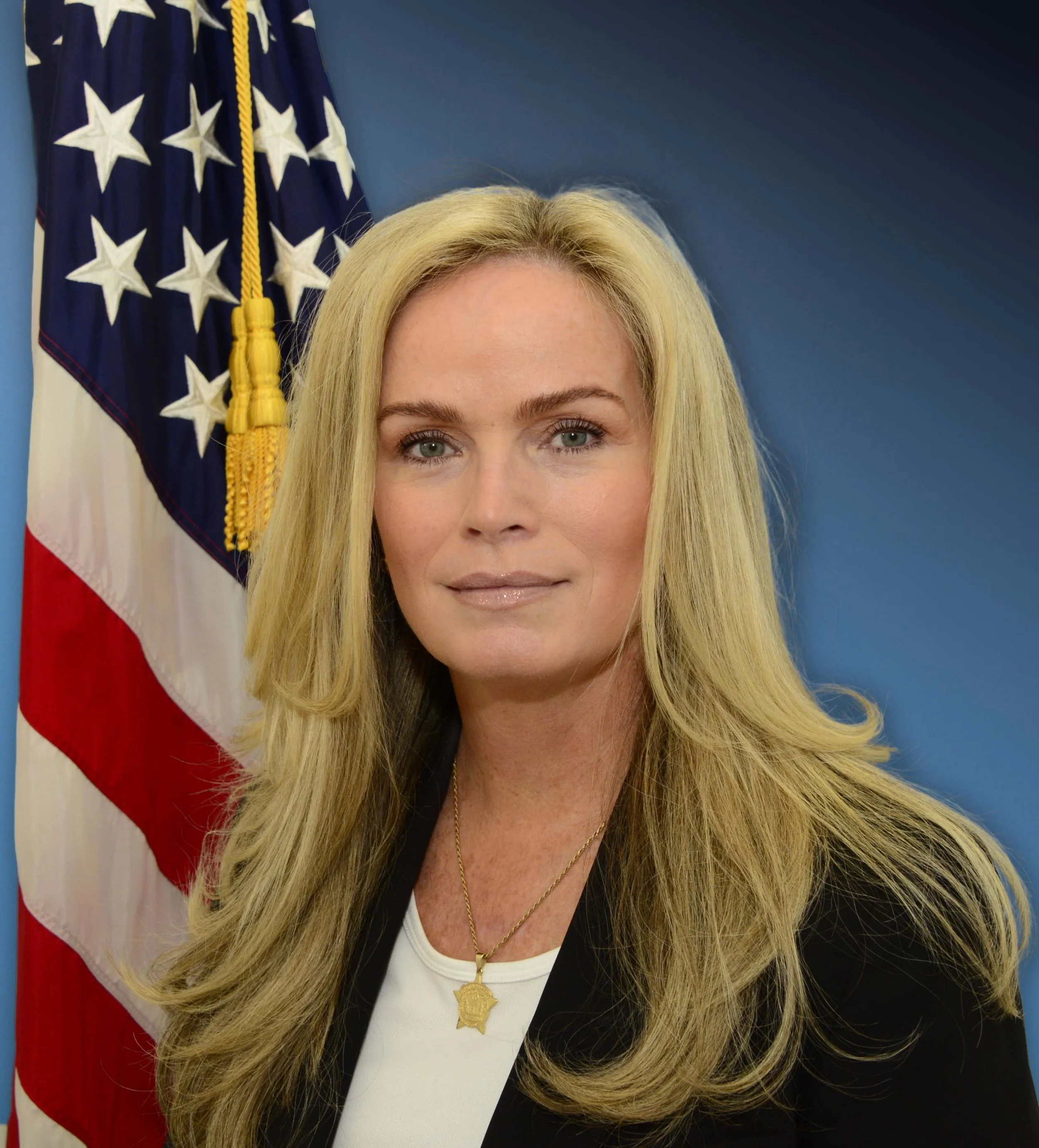 A woman with long blonde hair and green eyes standing in front of an American flag. She is wearing a black blazer, a white top, and gold jewelry, including a necklace with a pendant and a necklace with a badge symbol. The background features a blue wall.