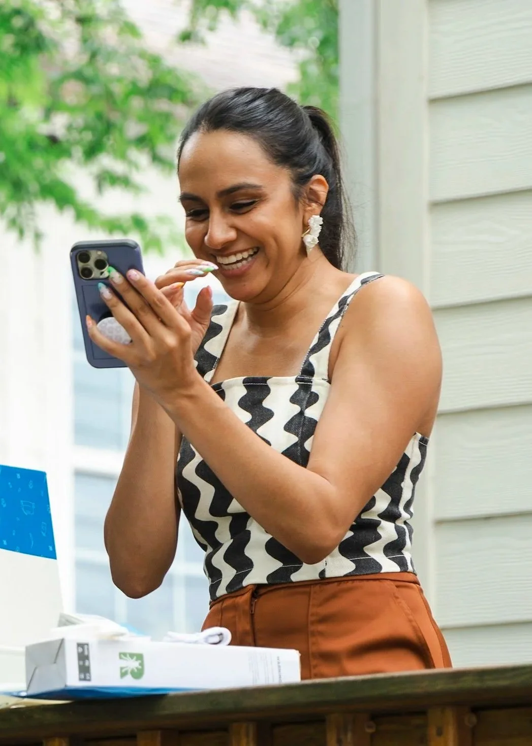 A woman with dark hair in a ponytail, wearing a black and white zigzag patterned sleeveless top and brown pants, is smiling and looking at her phone while standing outdoors near a house with horizontal siding and green trees in the background.