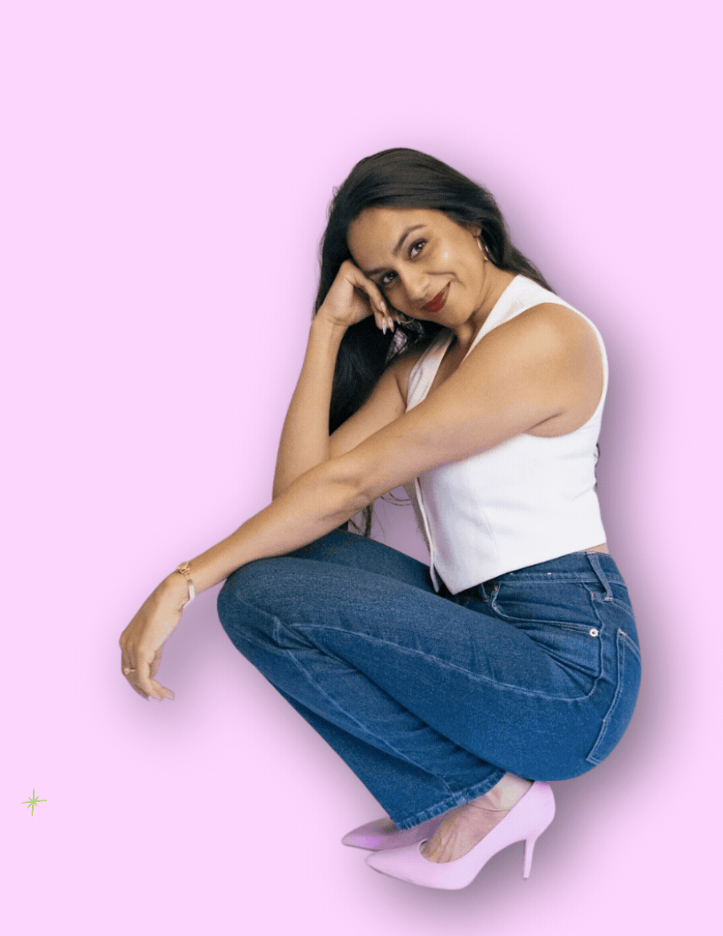 A woman with long dark hair, wearing a white sleeveless top, blue jeans, and pink high heels, squatting against a pink background, with her head resting on her hand and smiling.