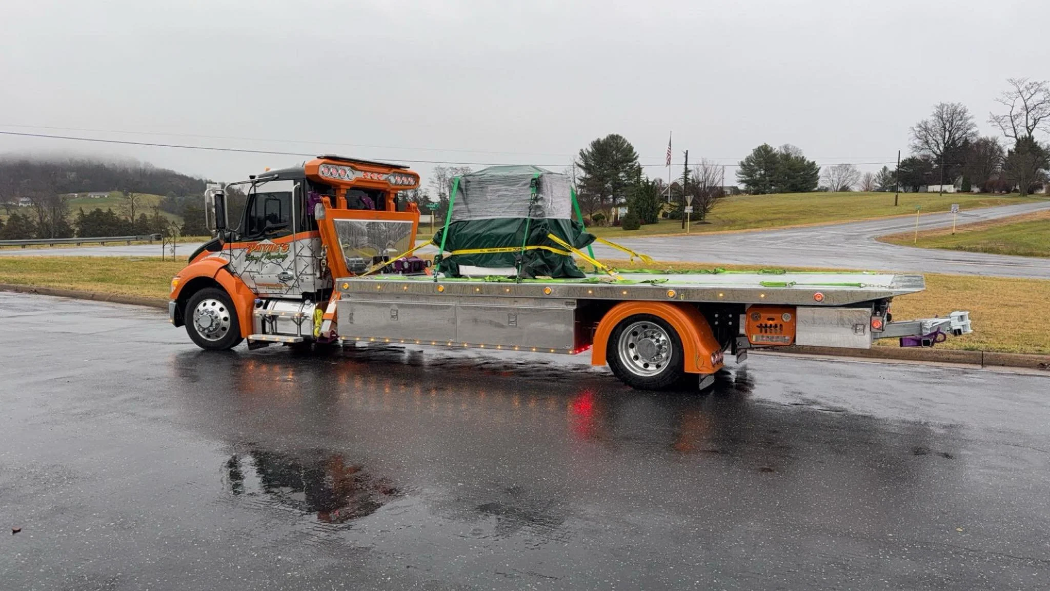 Orange tow truck parked on a wet asphalt road on a rainy day, with a wrapped and secured load on its flatbed, in a rural area with grassy fields, small trees, and a cloudy sky in the background.