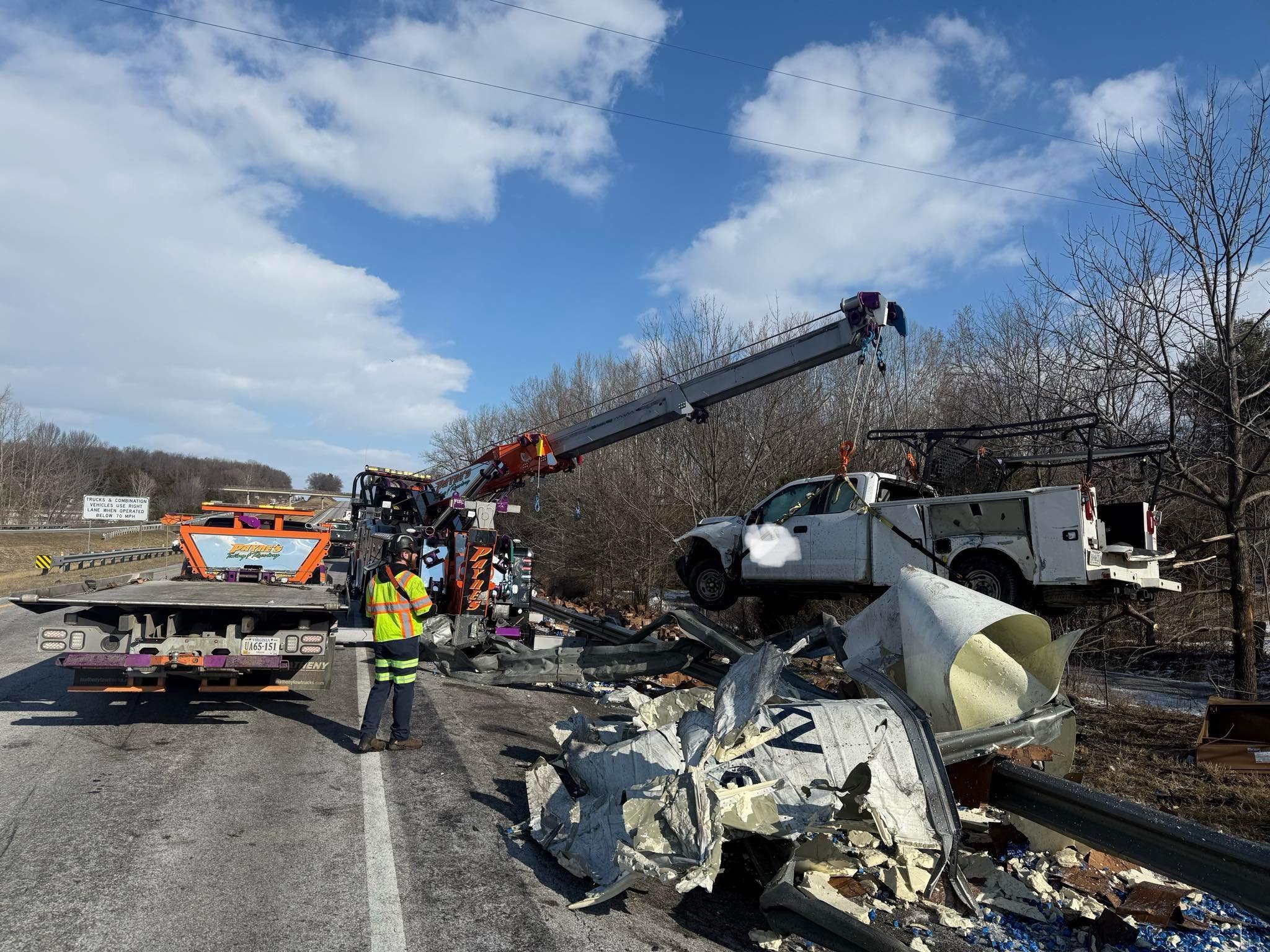 Accident scene on the highway with a damaged white pickup truck suspended by a crane, debris scattered on the road, and a worker in a yellow safety vest inspecting the wreckage under a partly cloudy sky.