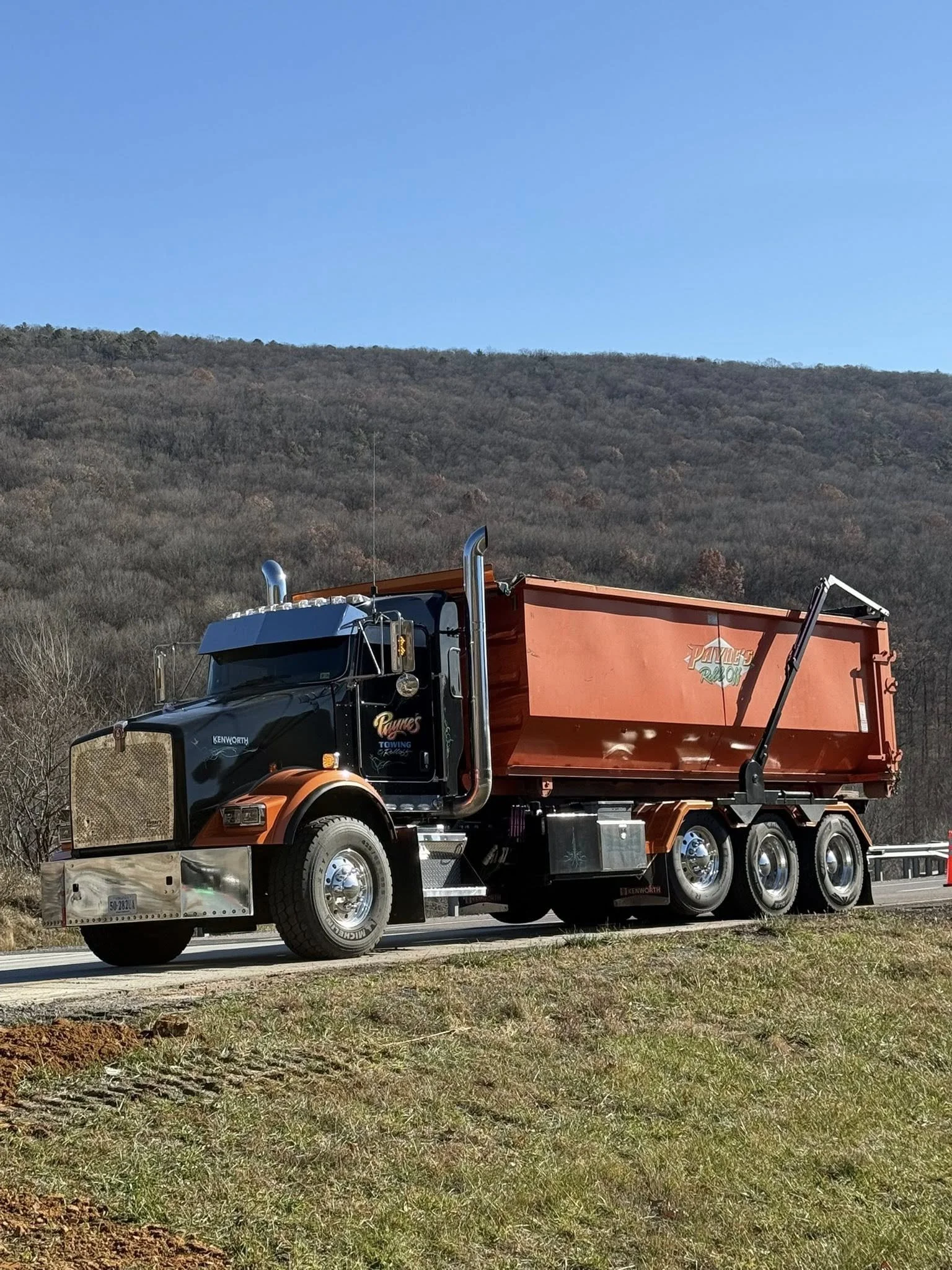 A large black and orange dump truck with the words 'Payne's Recycling' on the side, driving on a rural road with a wooded hillside in the background and a clear blue sky overhead.