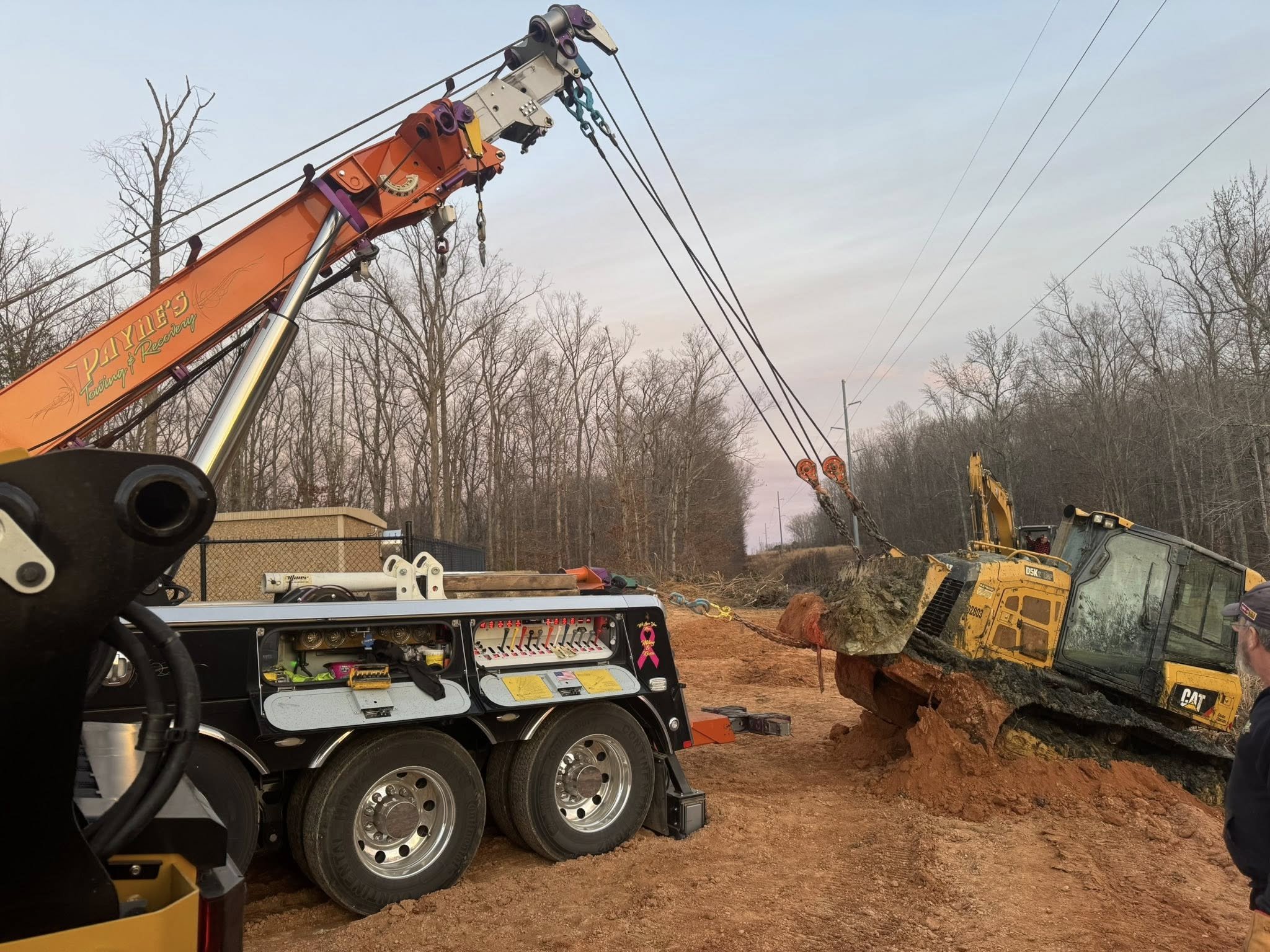 A construction scene with a crane lifting a large rock and a construction vehicle positioned on a dirt slope, surrounded by leafless trees and power lines in the background.