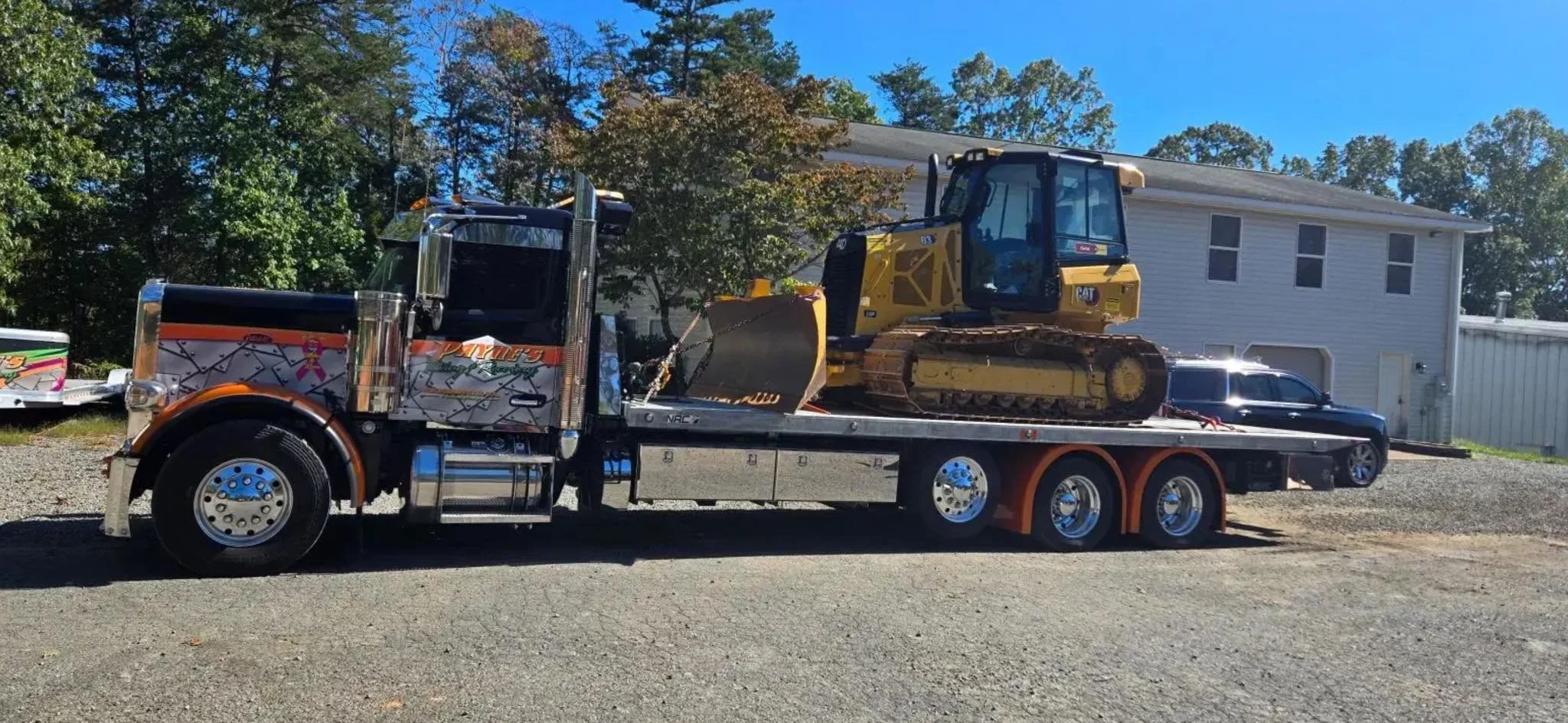 A large black tow truck with chrome accents transporting a yellow CAT excavator with a black cab on its flatbed. The scene is outdoors with trees and a white building in the background on a sunny day.